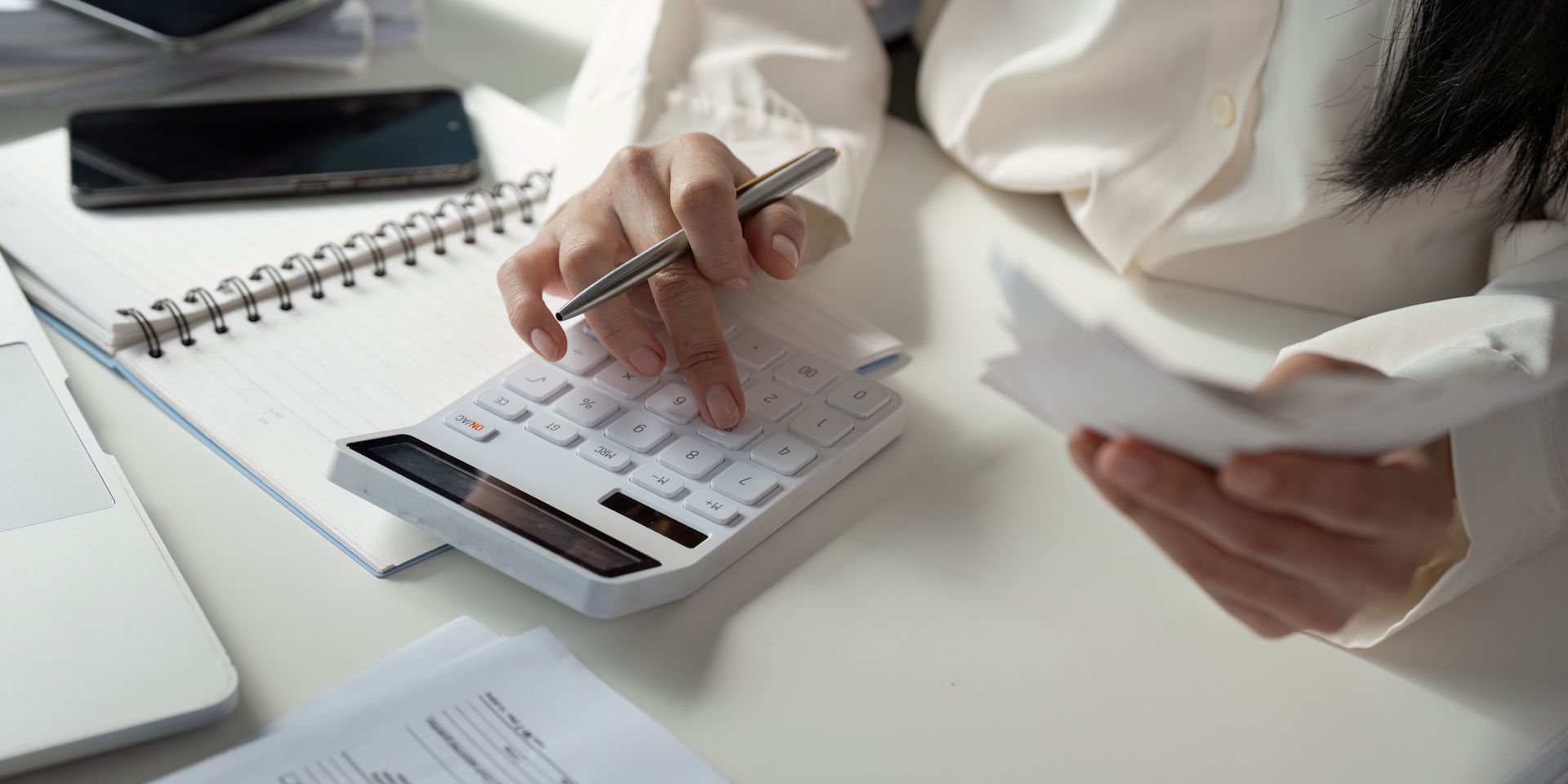 Person using a calculator while holding papers, possibly reviewing financial documents at a desk.