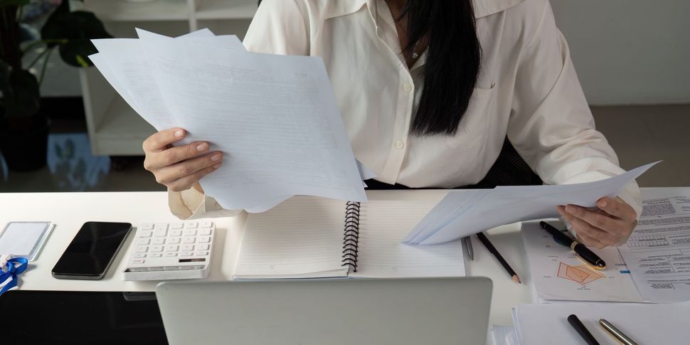 Person reviewing paperwork at a desk with a calculator, phone, and laptop.