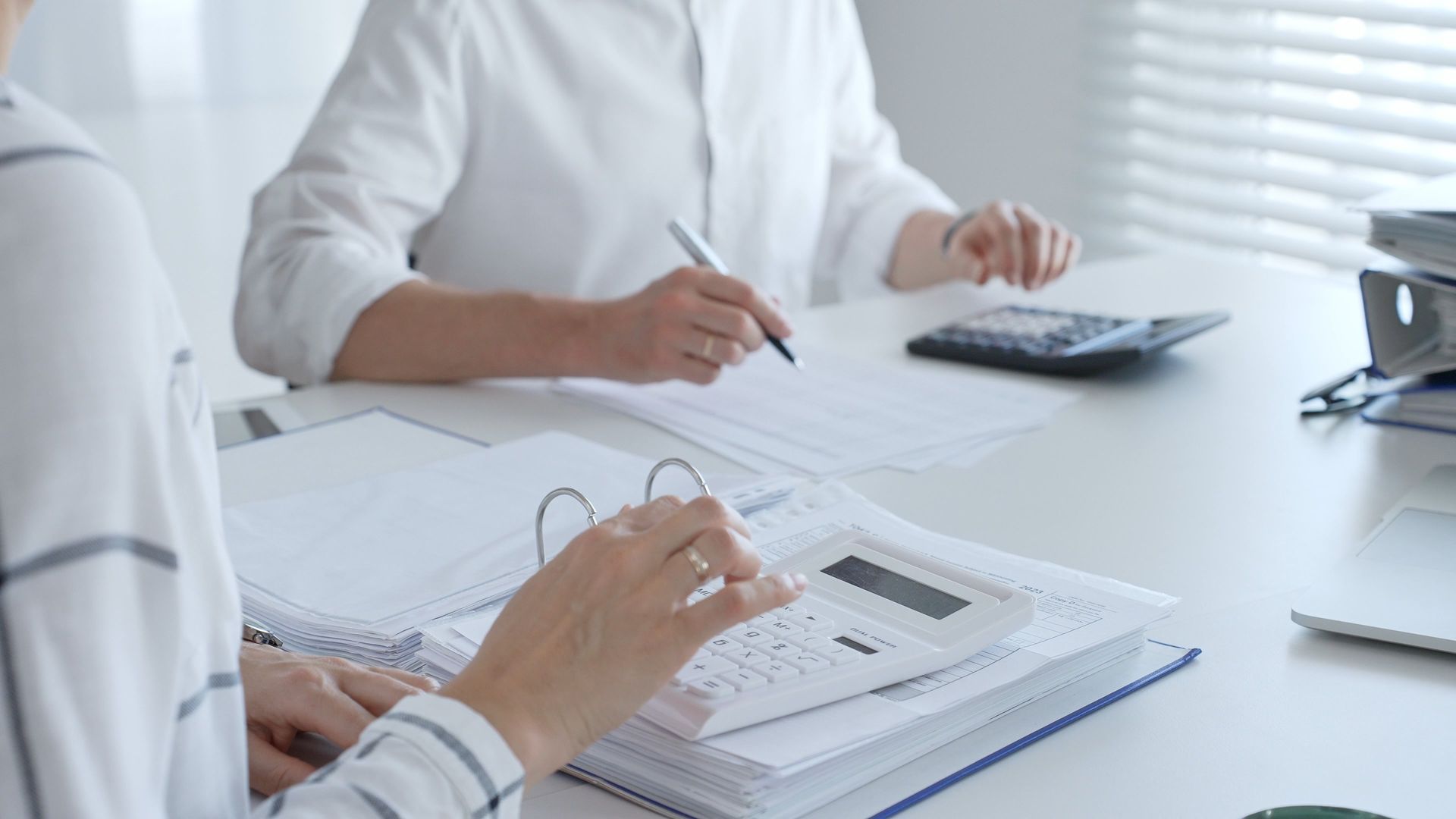 Two people calculating finances at a desk with papers, binders, and calculators in an office setting.