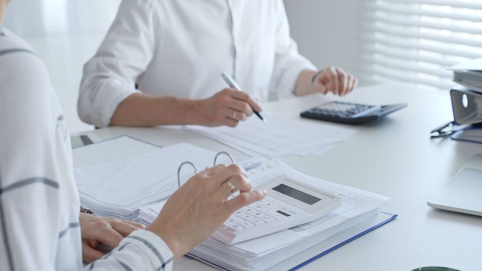 Two people calculating finances at a desk with papers, binders, and calculators in an office setting.