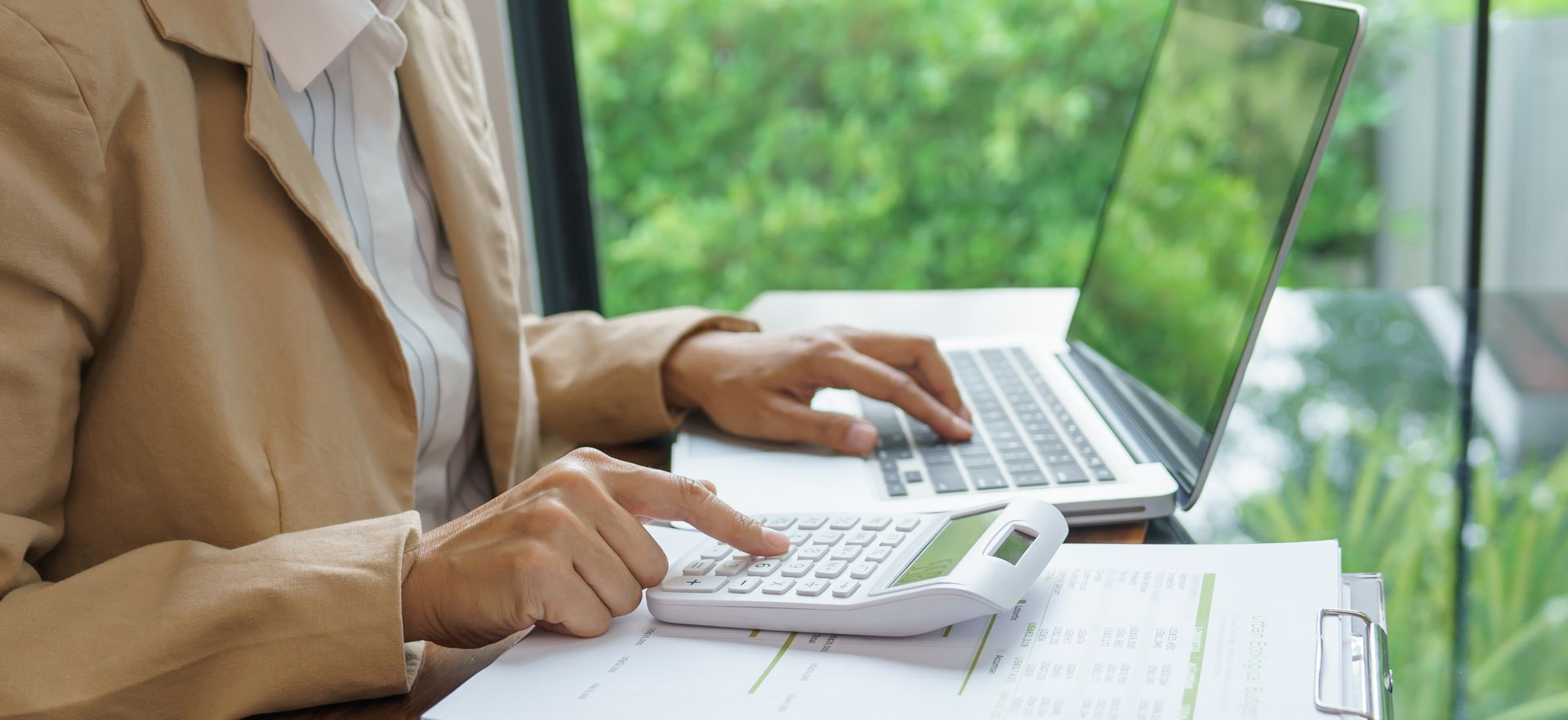 Person using a calculator and laptop, working at a desk with paperwork, near a window with a green view.