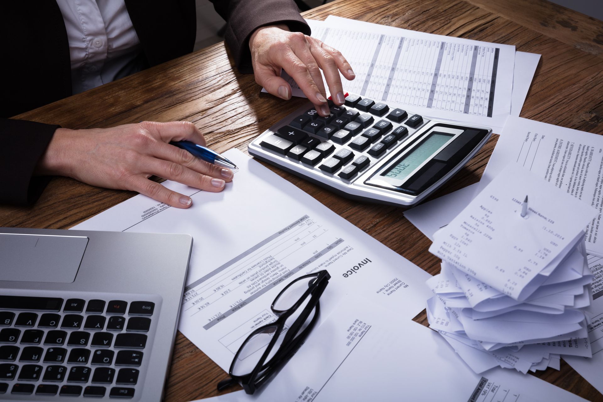 Hands using calculator, surrounded by papers, laptop, glasses on a wooden desk.