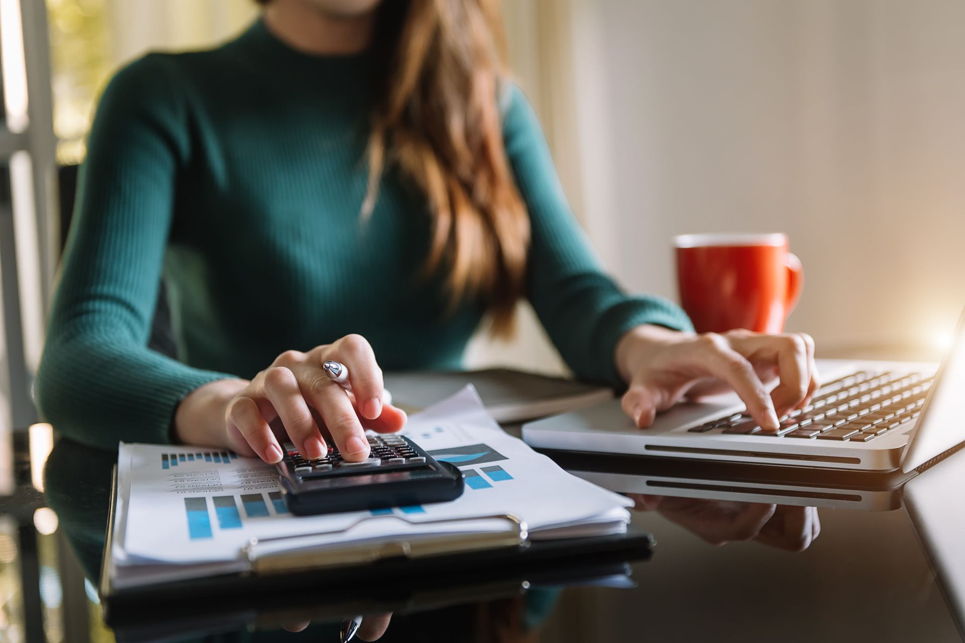 A person in a green sweater works at a desk with a laptop, calculator, and financial charts, with a red mug nearby. A person in a green sweater works at a desk with a laptop, calculator, and financial charts, with a red mug nearby.