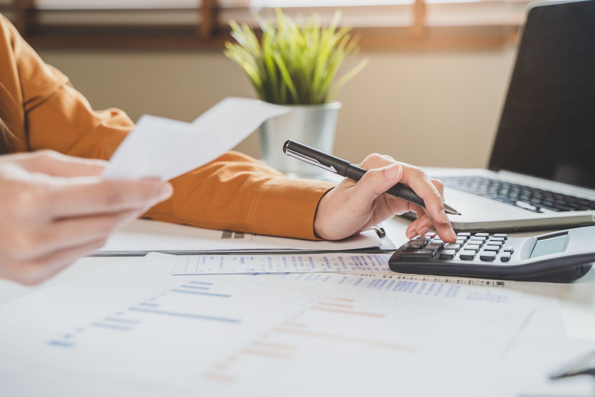 Person using a calculator, holding papers, and working on a laptop at a desk.