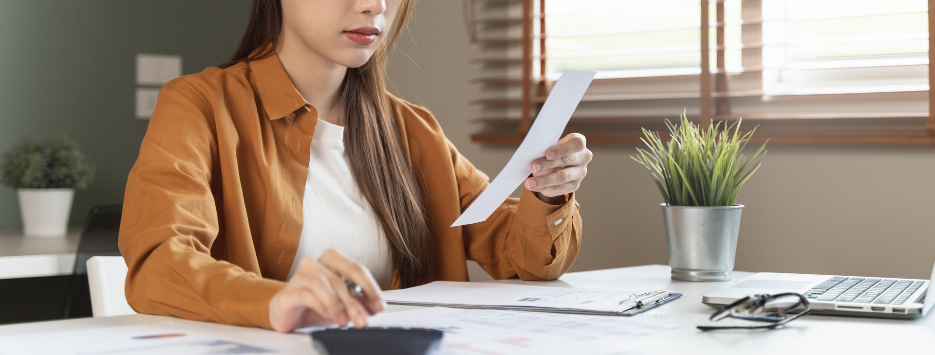 Woman reviews paperwork at a desk with calculator, laptop, and plant.