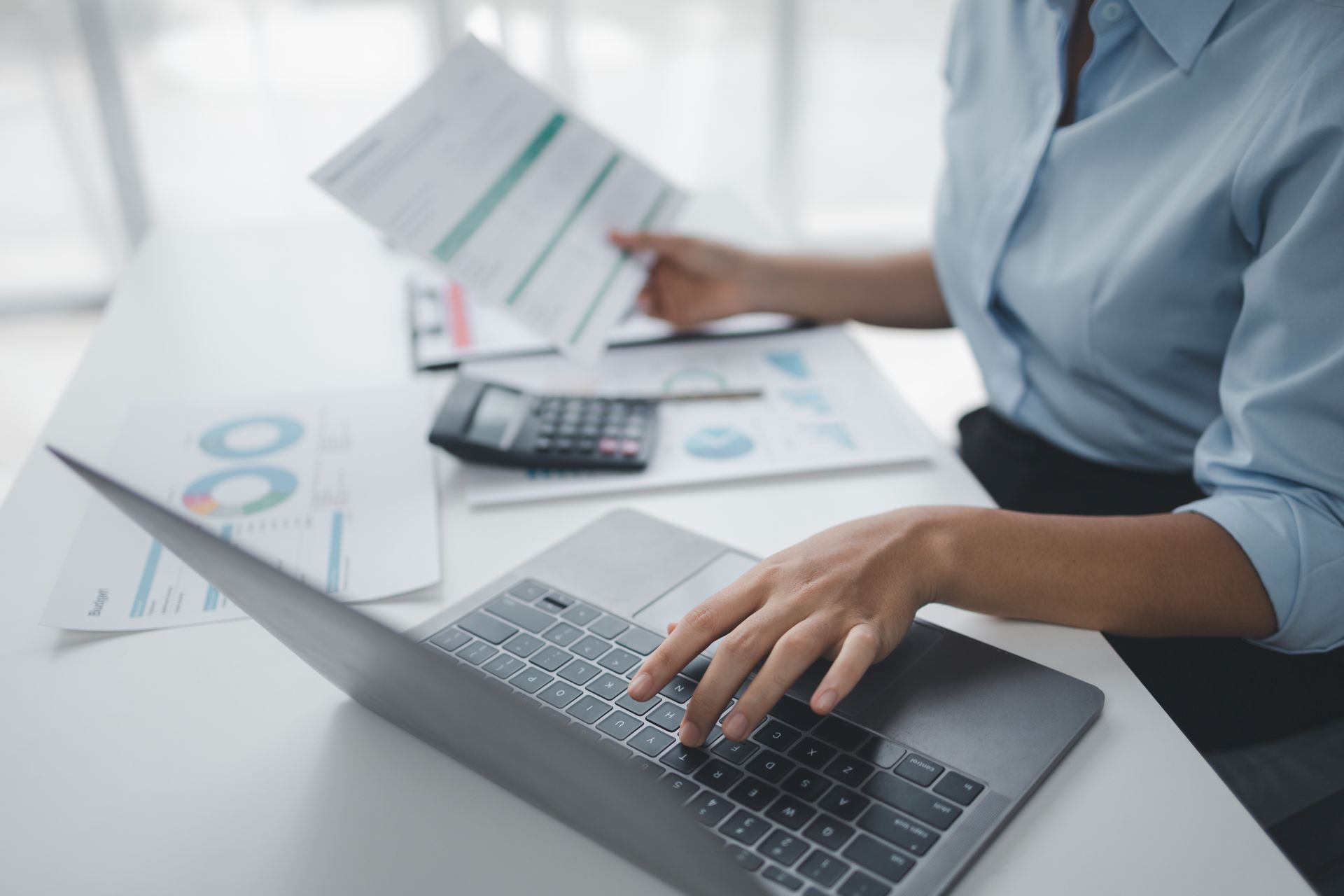 Person working on laptop, reviewing documents, and using a calculator at a desk.