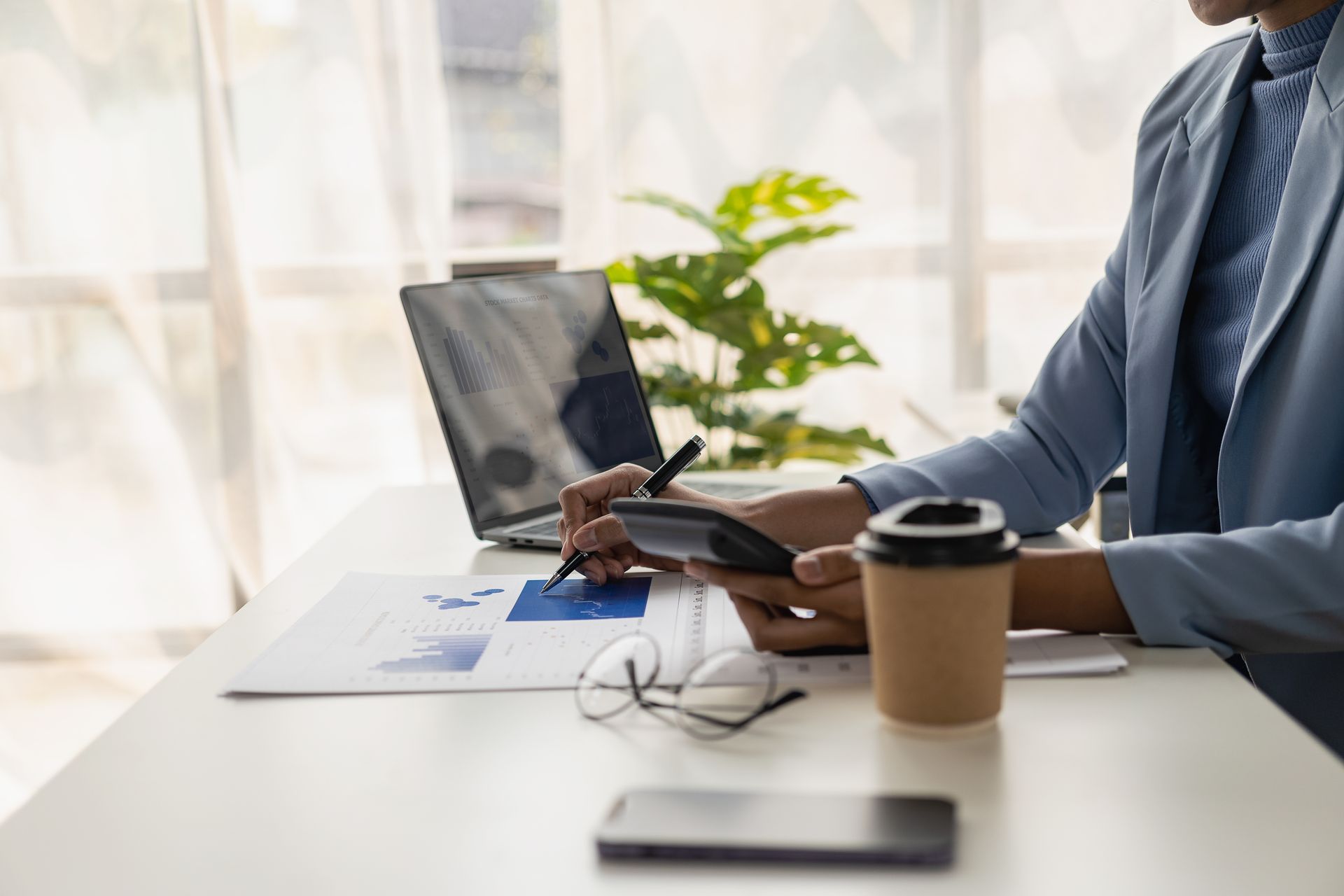 Person in blue blazer, calculating with a calculator, reviewing charts at a desk with a laptop and coffee.