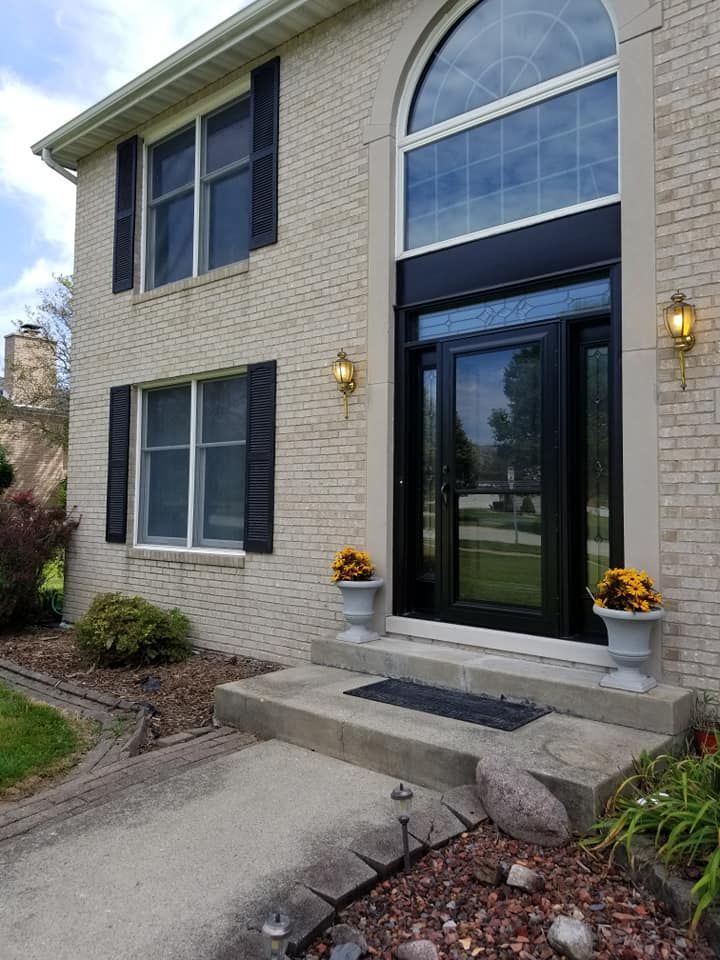 Beige brick house with black front door, shutters, and arched window. Potted flowers flank the entry.