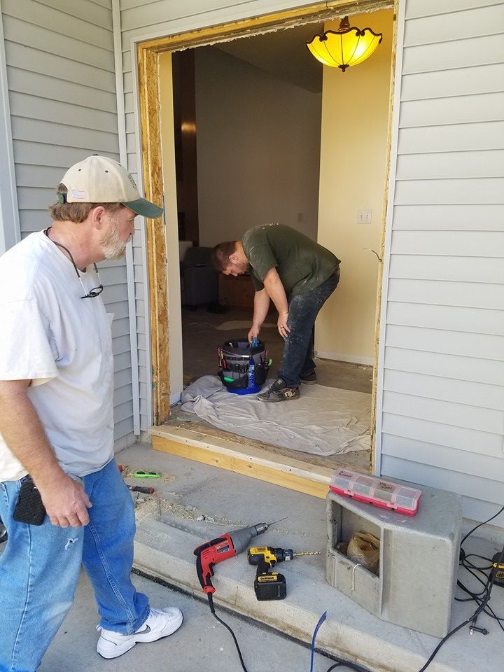 Two men work on a doorway repair. One observes, the other bends over tools. Outdoor setting with a porch.
