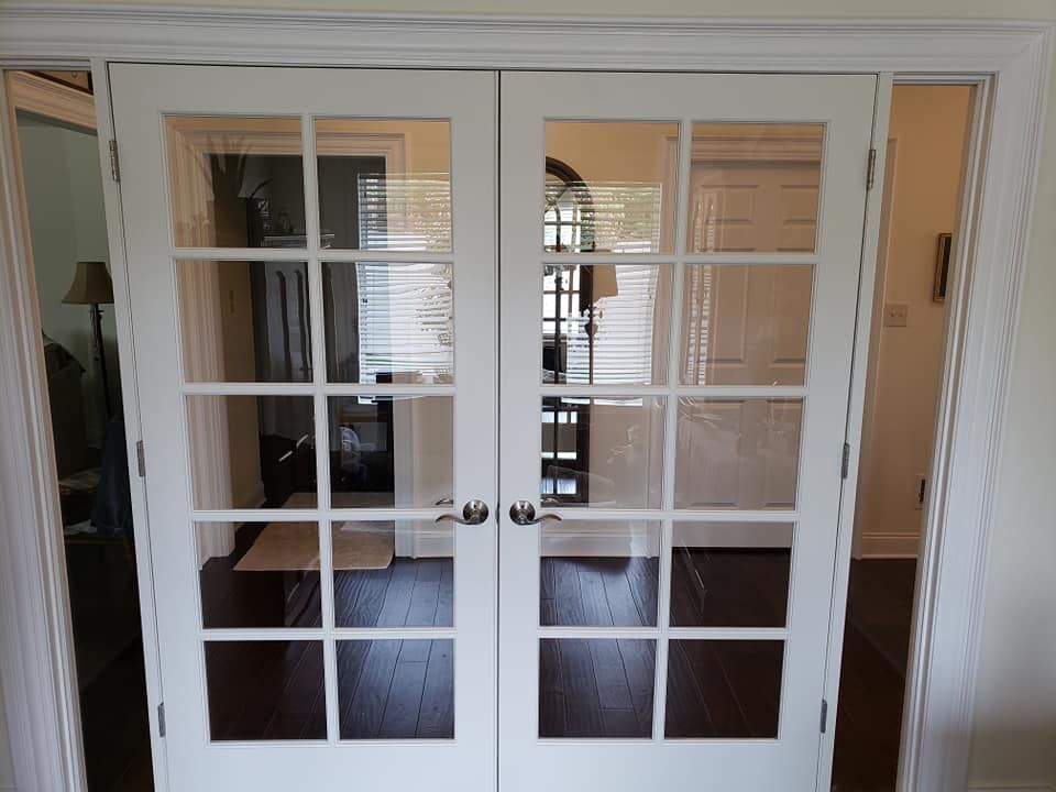 White-framed French doors with glass panes, reflecting a room with a table, blinds, and dark wood flooring.