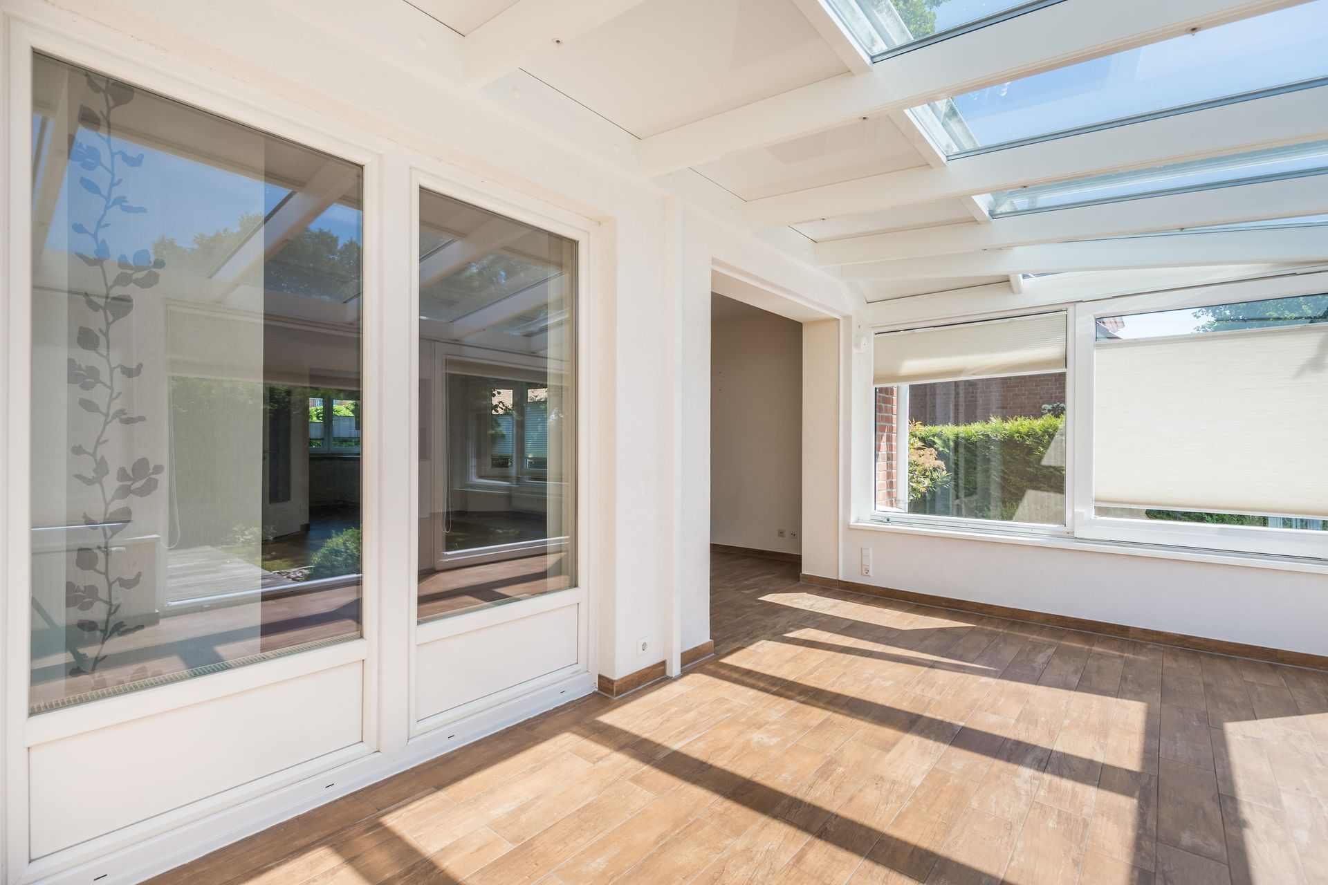 Sunroom with glass walls and roof, wooden floor, white trim. Sunlight streams in.