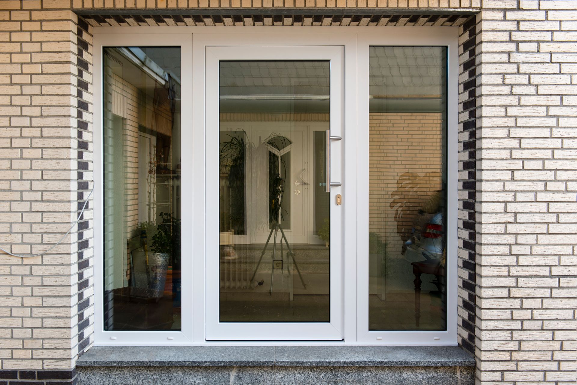 White-framed glass entry door with side panels, set in a brick wall. Reflects a bright interior and tripod.