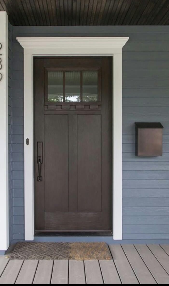 Dark wood Craftsman-style front door with a six-pane window and white decorative trim, set against blue siding with a bronze mailbox.