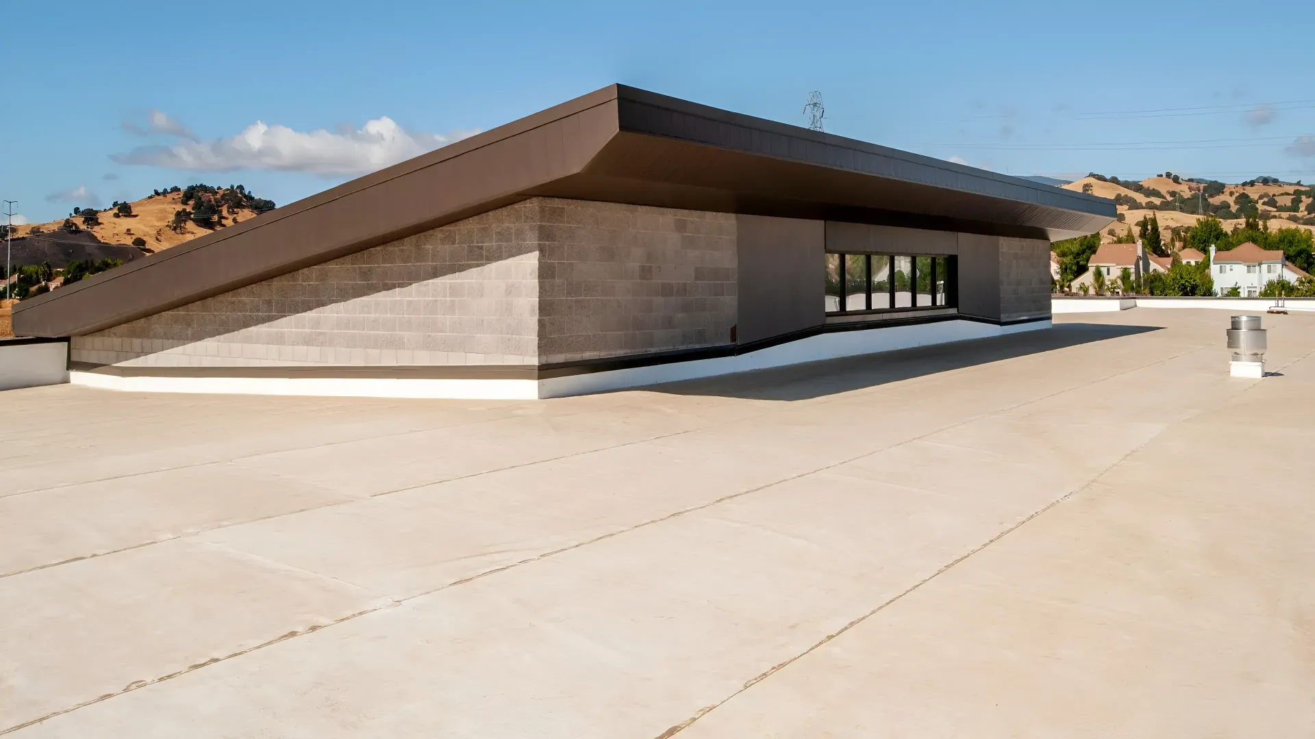 A modern building with a brown sloped roof over a flat, light-colored roof. Rolling hills in the distance.