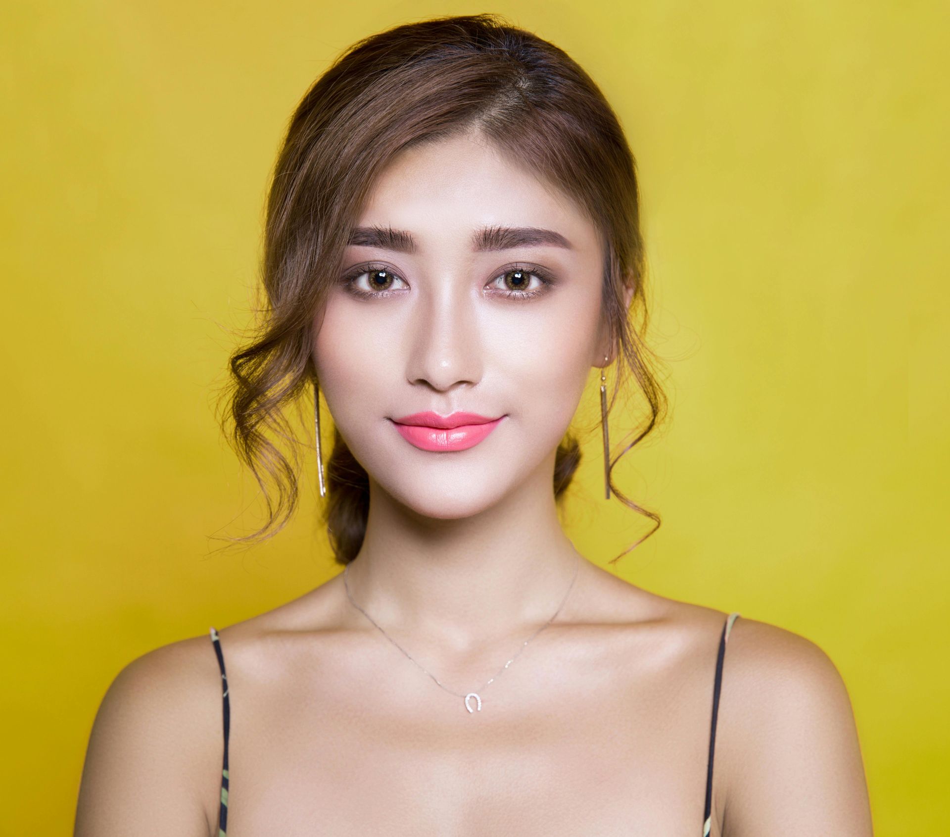 A woman wearing a necklace and earrings is standing in front of a yellow background.