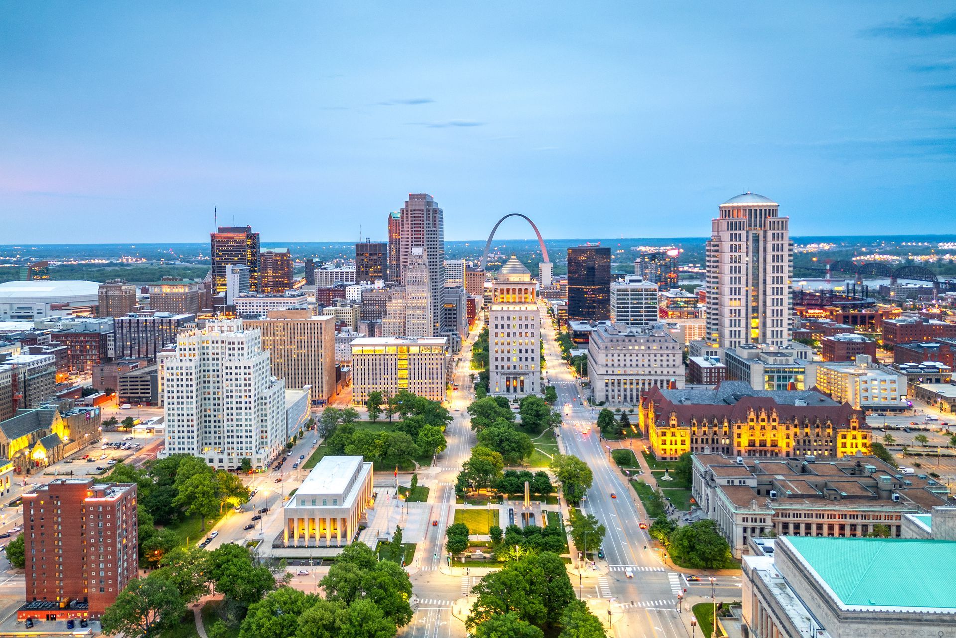 Aerial view of St. Louis skyline at dusk, with the Gateway Arch in the distance and a tree-lined boulevard.