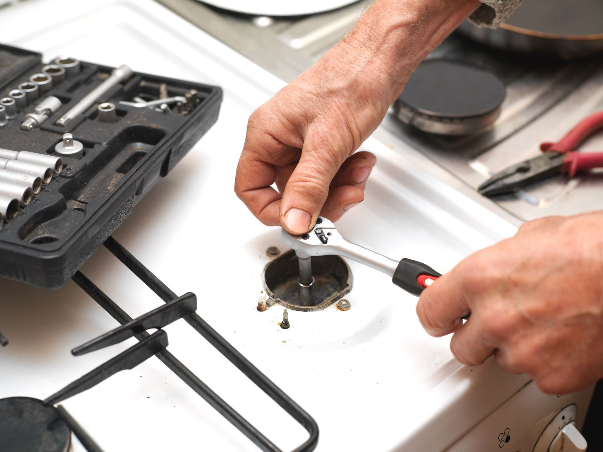 Person using a wrench to repair a stovetop, with a toolbox and pliers nearby.