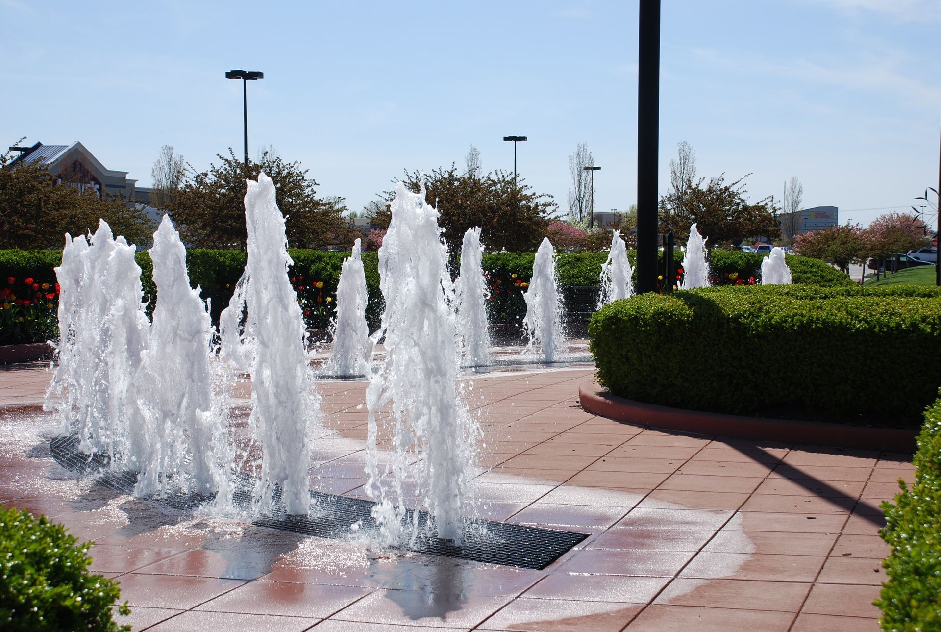 Fountains spraying water in a landscaped plaza with red brick and green bushes. Bright, sunny day.