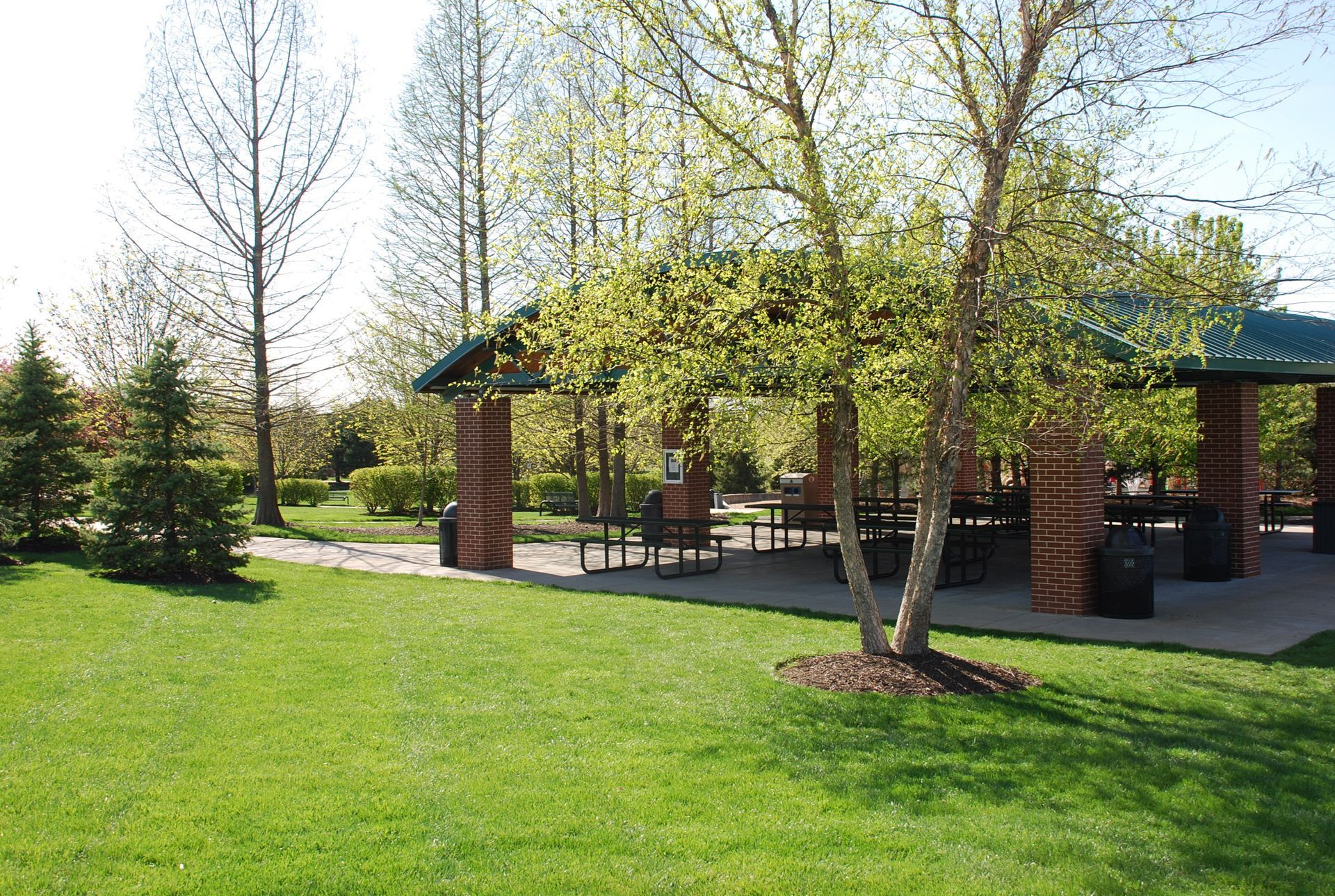 Park pavilion with picnic tables, surrounded by green grass and trees.