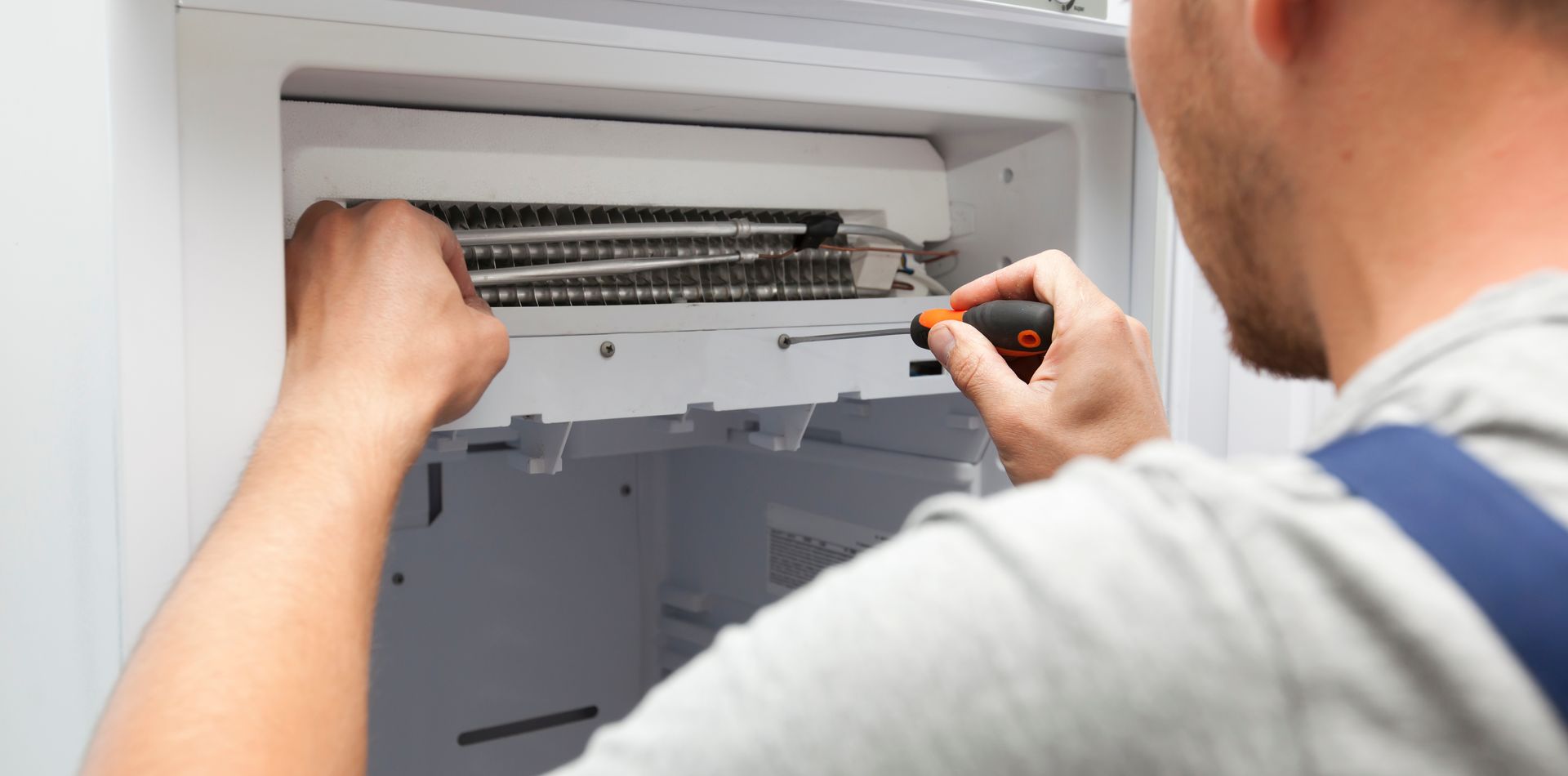 A person repairing a refrigerator with a screwdriver. The person's back is to the camera.