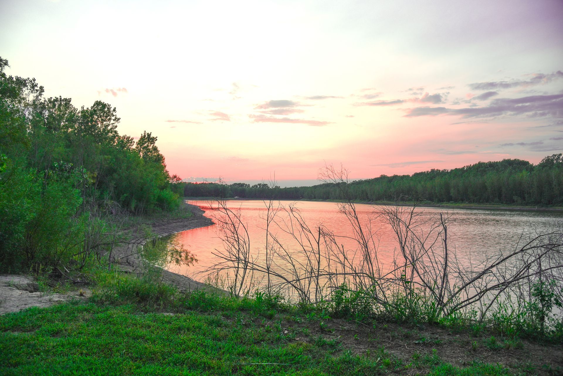 Sunset over a lake; orange and pink sky reflects on the water. Trees line the shore with green grass.