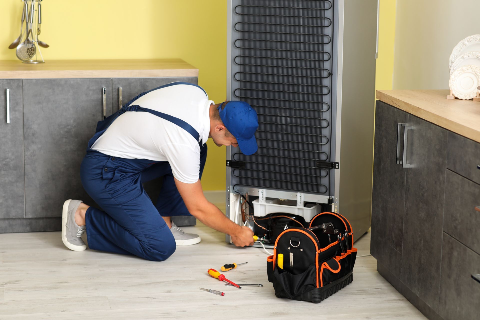 Person in blue overalls repairs a refrigerator in a kitchen, tool bag nearby.