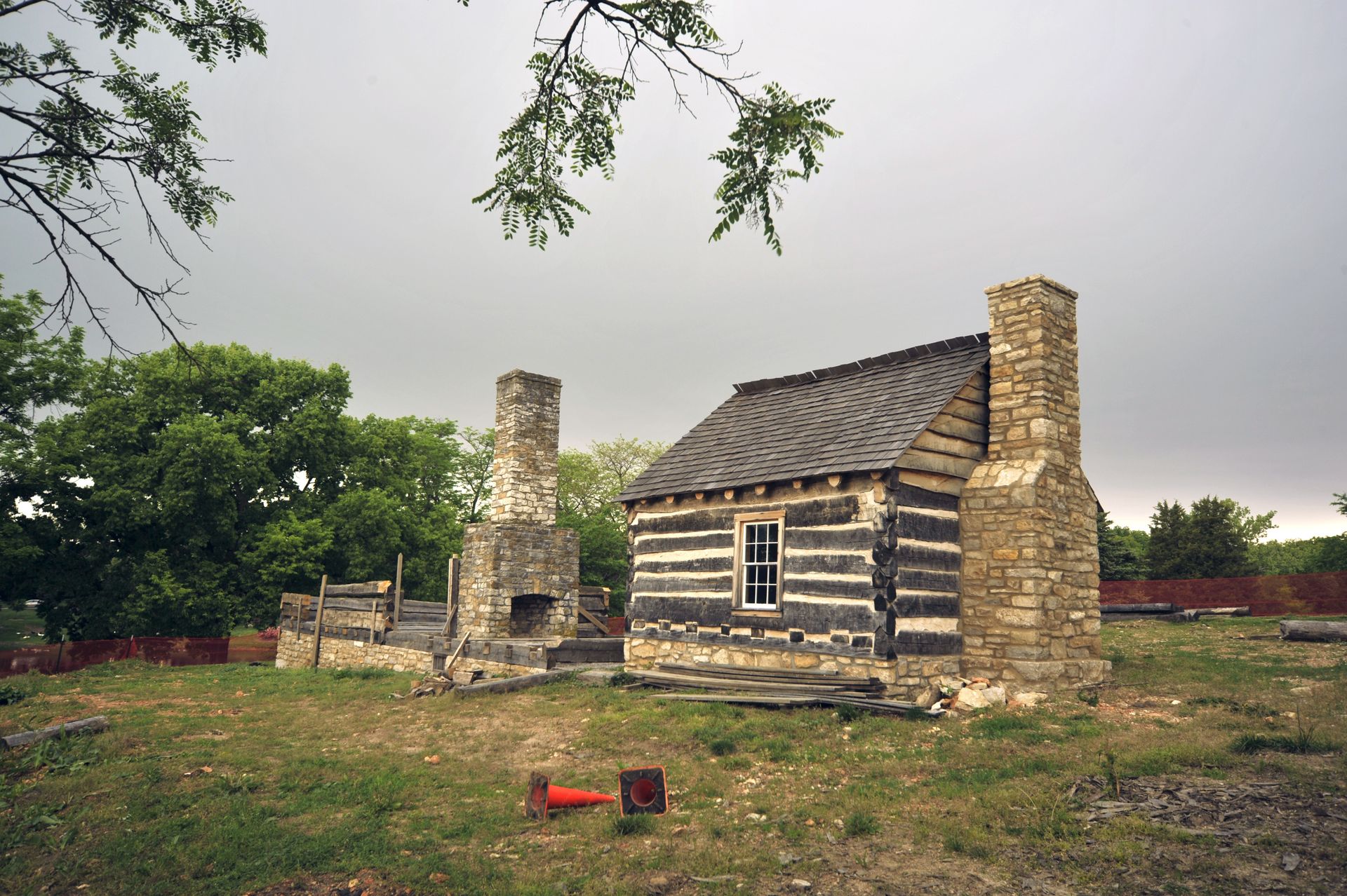 Log cabin with stone chimneys in a grassy field on a cloudy day.