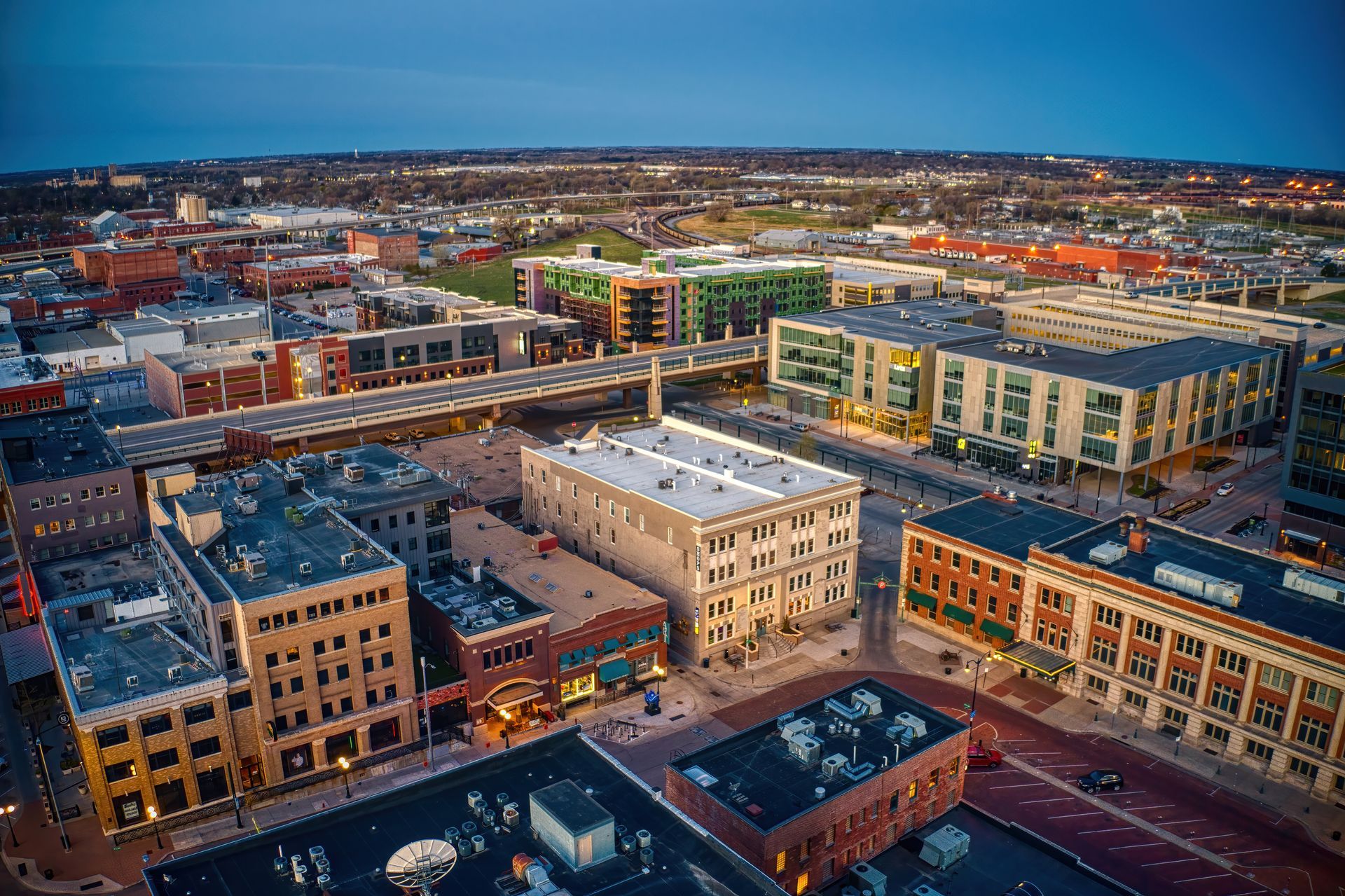 Aerial view of a city at dusk; buildings with lit windows under a twilight sky.