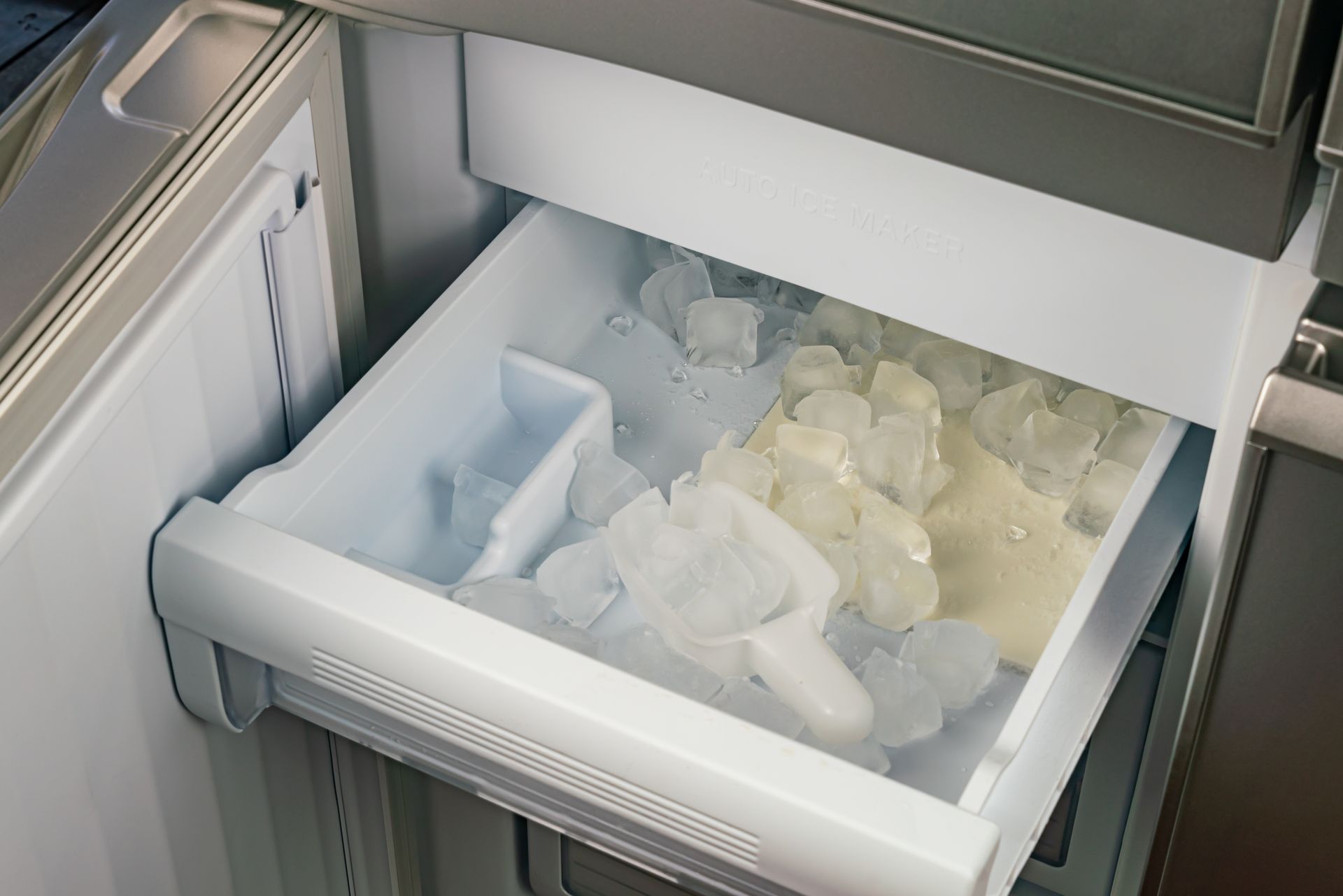 Open refrigerator drawer filled with ice cubes and scoop.