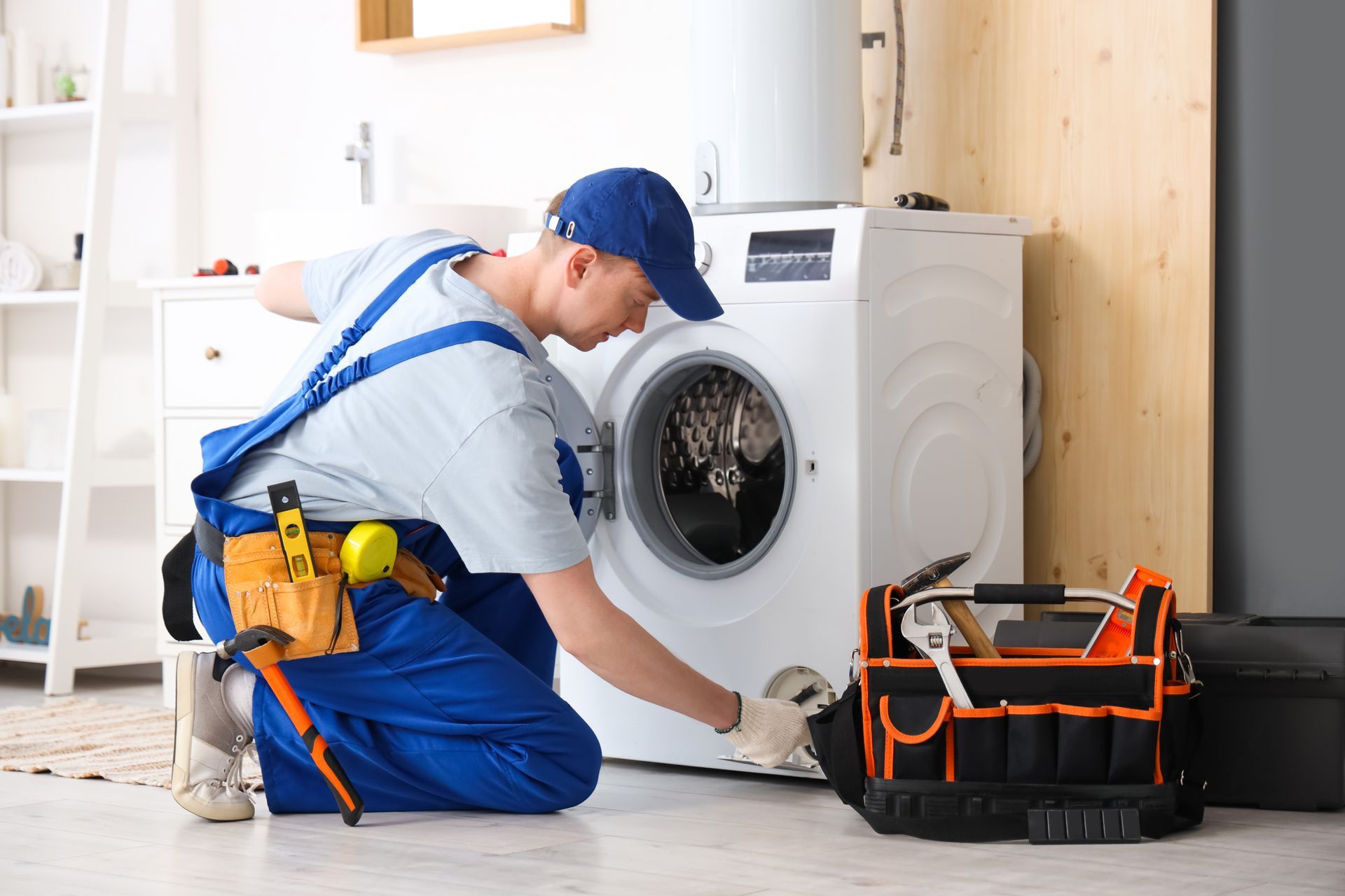 Man in blue overalls repairs a washing machine in a laundry room. Toolbox and tools on the floor.