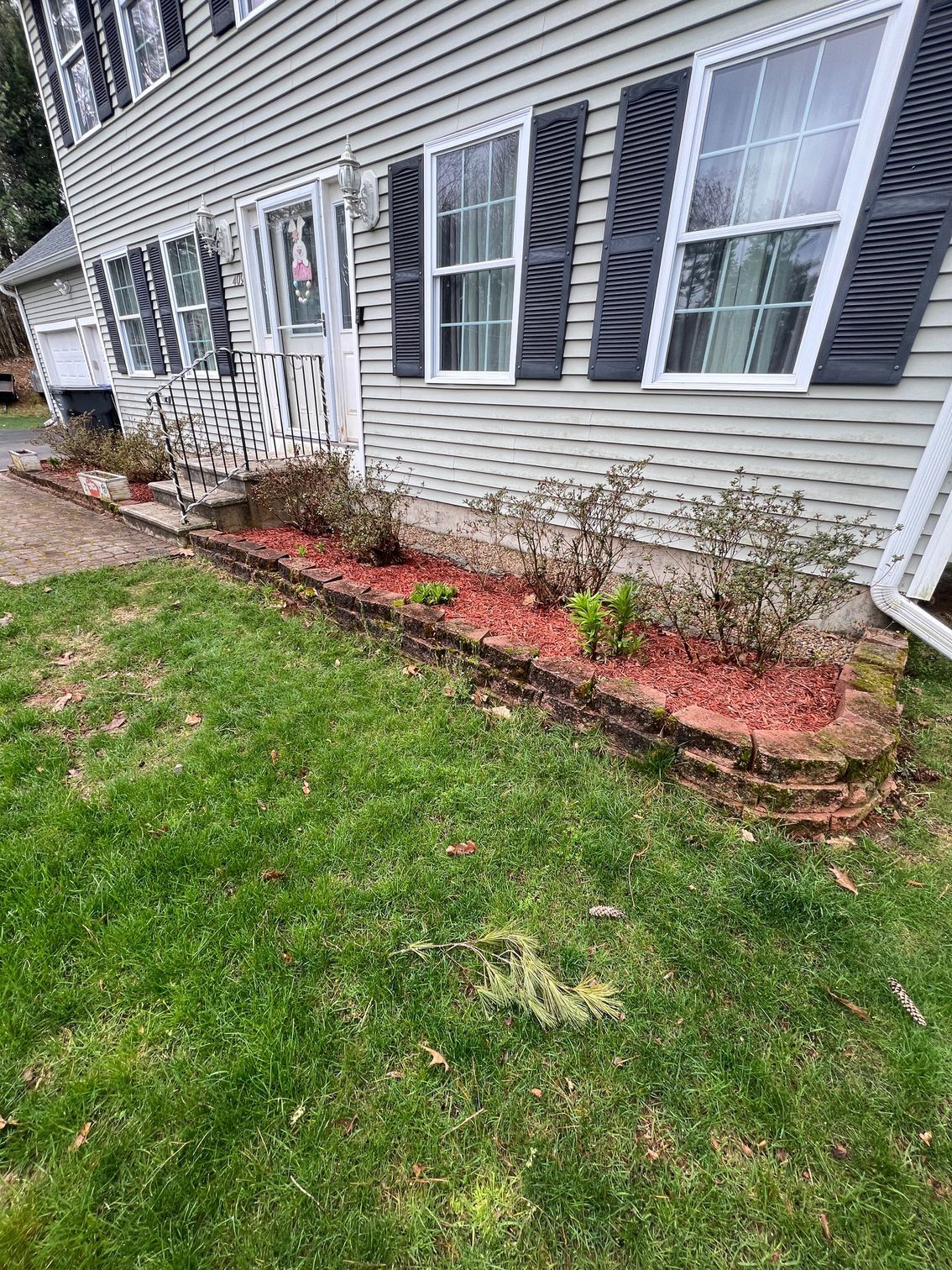 A house with gray siding and black shutters, featuring a red mulch flower bed in front, edged with brick, with a green lawn.