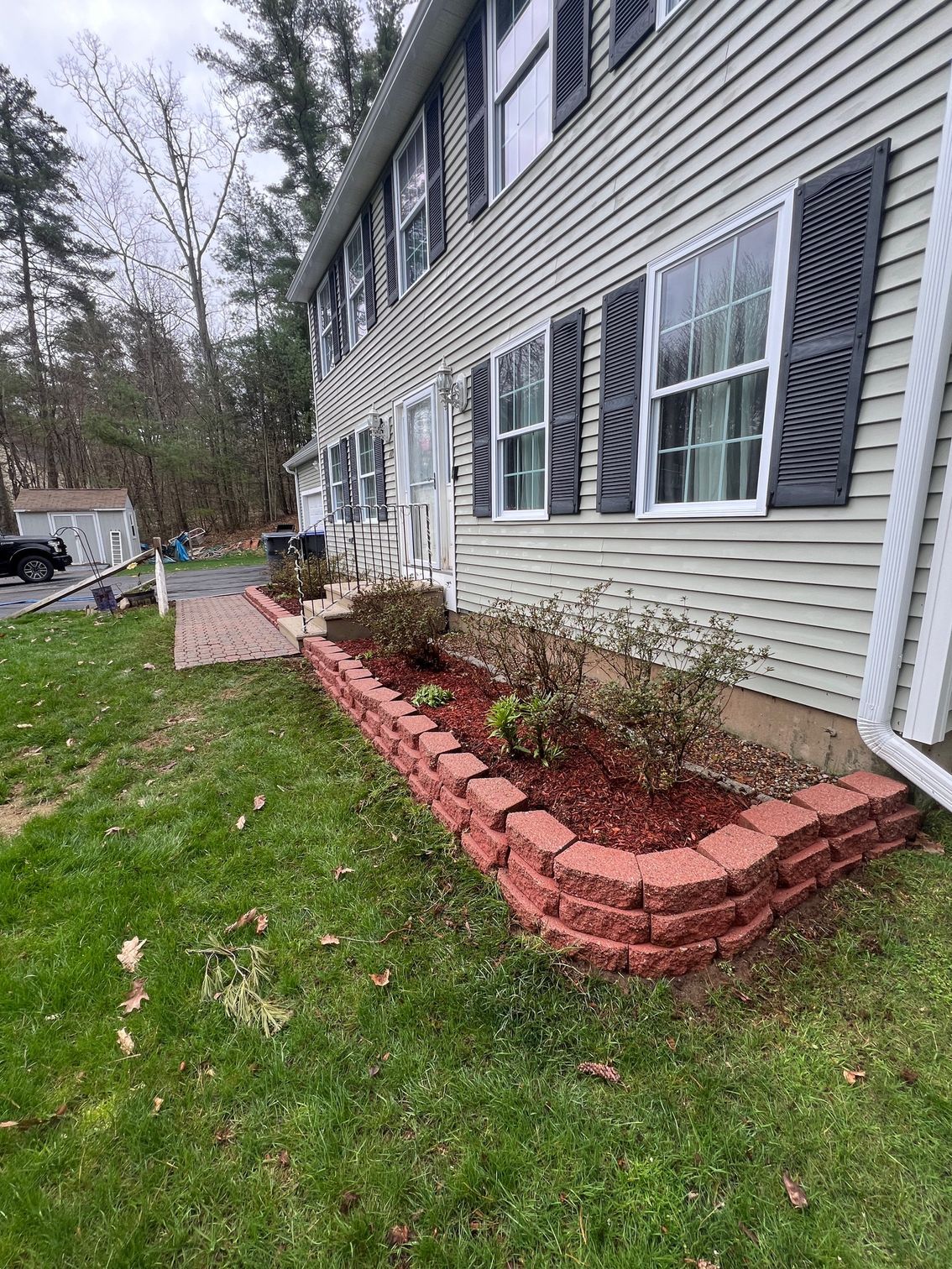 A two-story house with a red brick retaining wall and flower bed, alongside a walkway. Green grass surrounds the house.