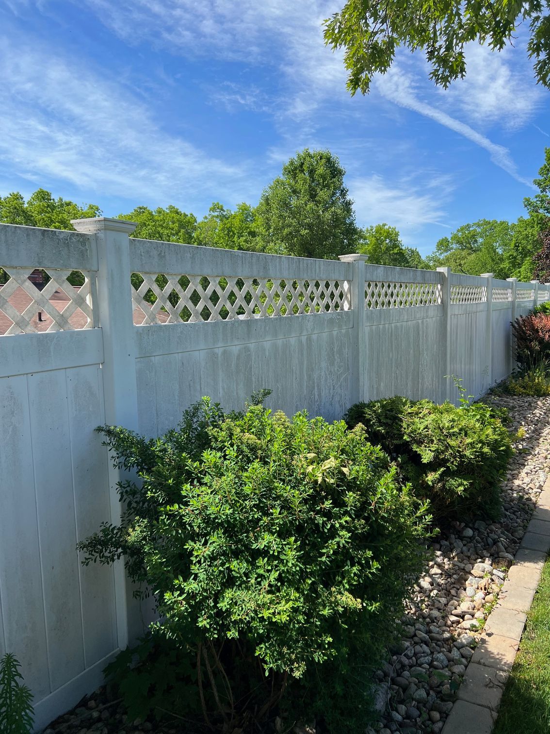 White fence with decorative lattice panels, green bushes in front, and a bright blue sky with clouds.