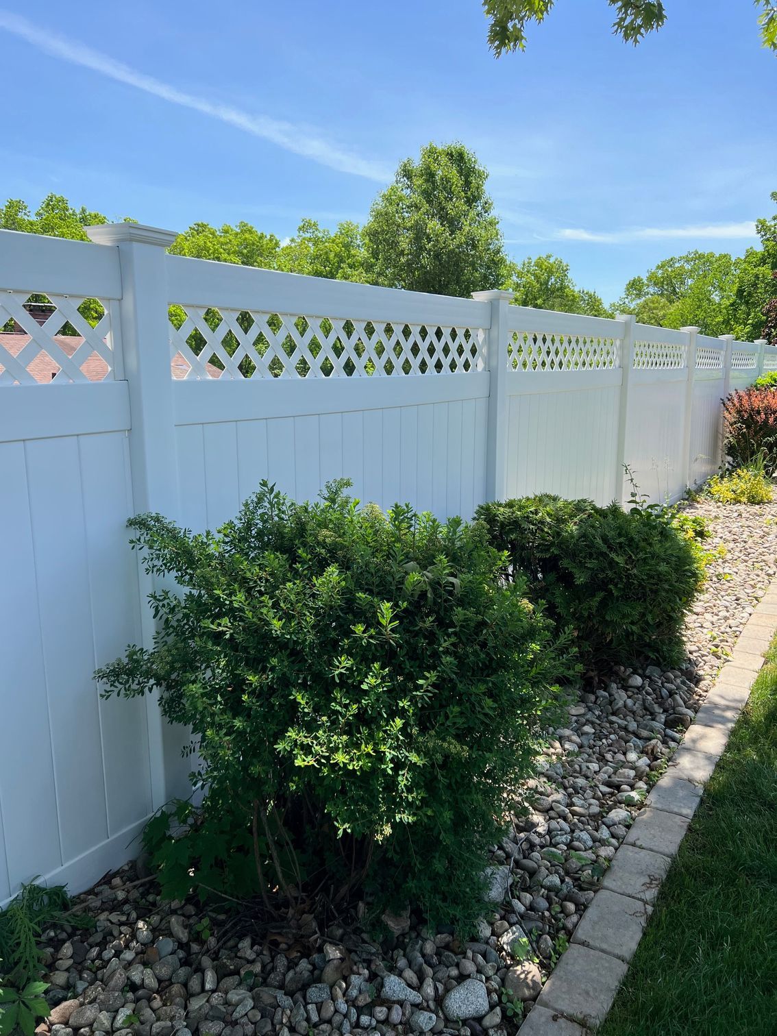 White vinyl fence with lattice detail on a sunny day; green bushes and a rocky bed border the fence.