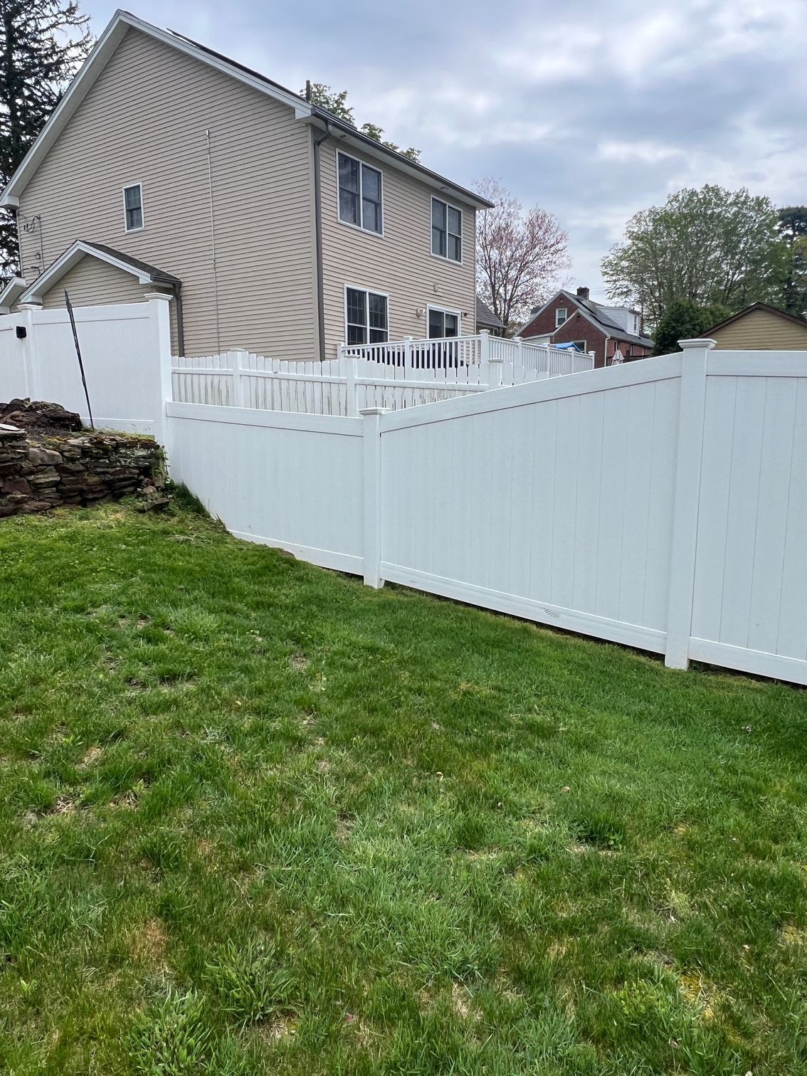 White vinyl fence surrounding a grassy backyard, with a two-story beige house in the background. The sky is overcast.