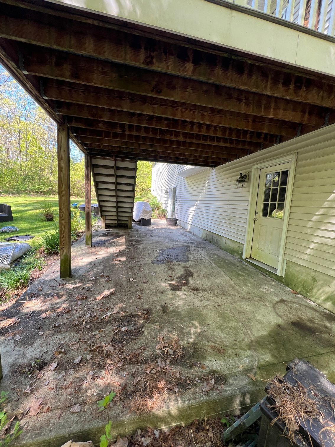Exterior view under a wooden deck with a concrete patio, next to a white house with a door.