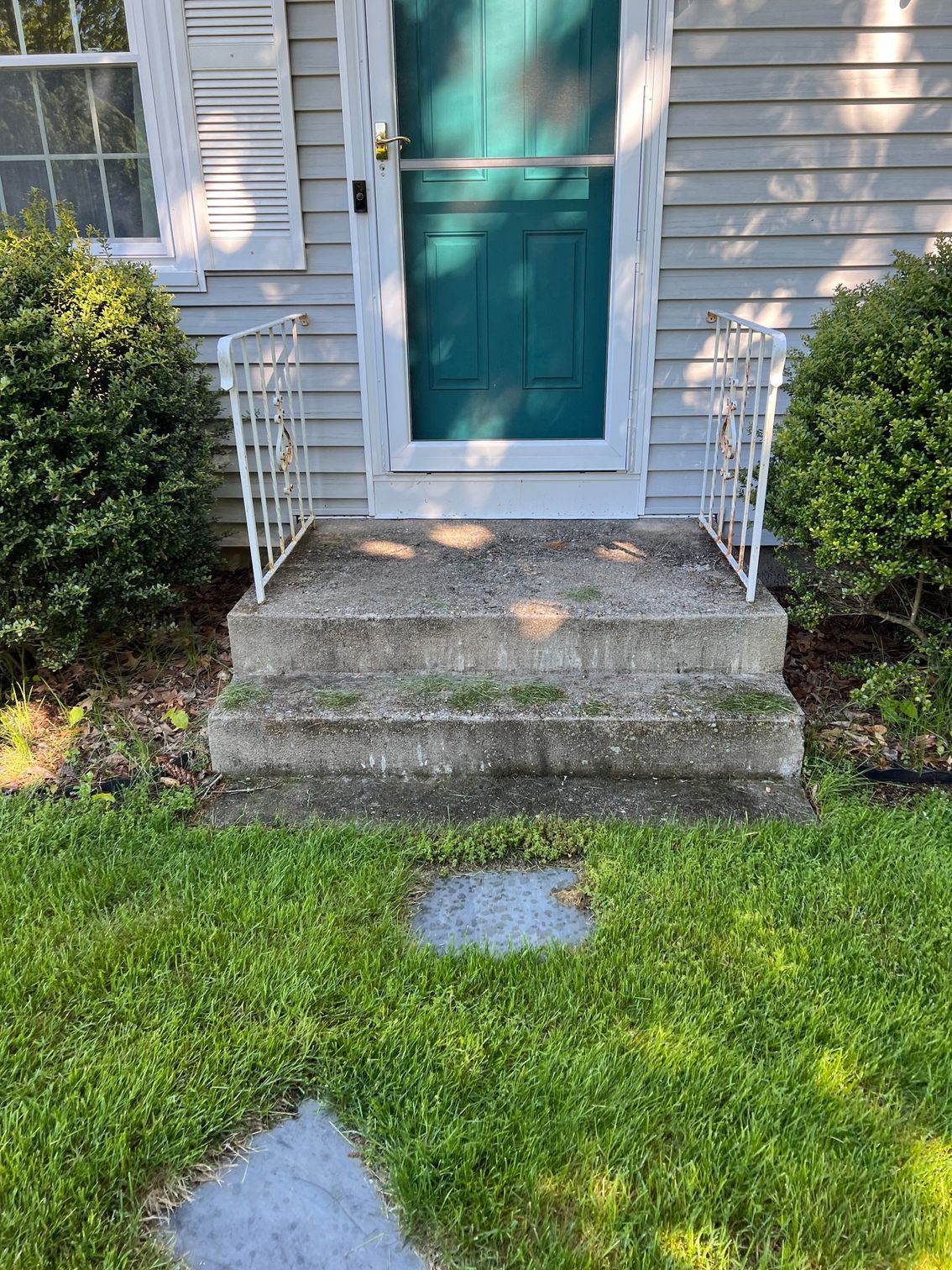 Concrete steps leading to a teal door with white railing and bushes.