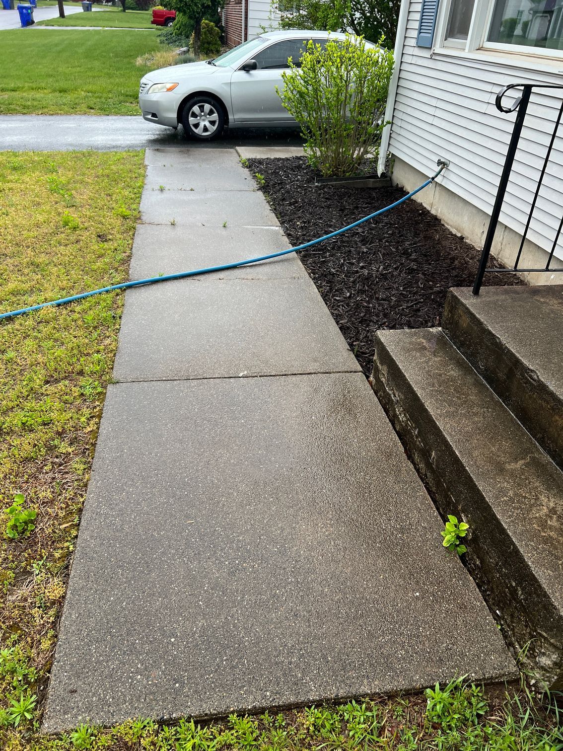 Concrete sidewalk leading to steps of a white house, blue hose across it. A car is parked in the background.