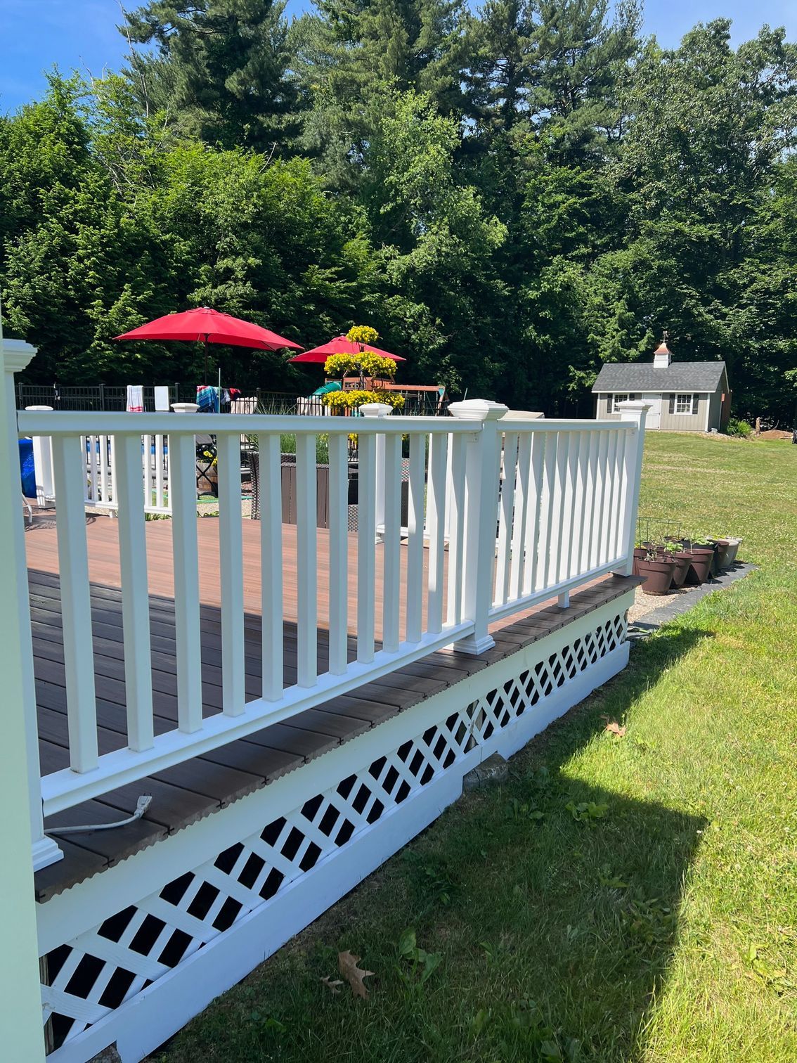 White picket fence surrounds a brick patio with red umbrellas, overlooking a grassy yard with trees.