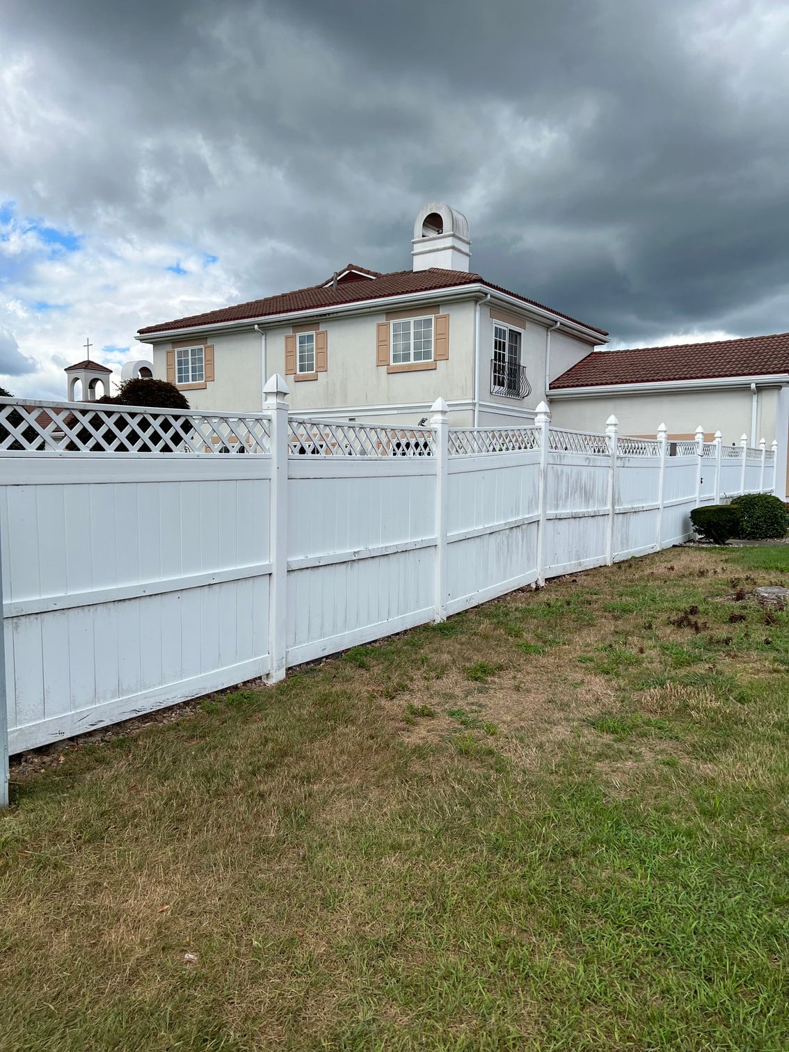 White vinyl fence in front of a house with a brown roof, on a cloudy day.