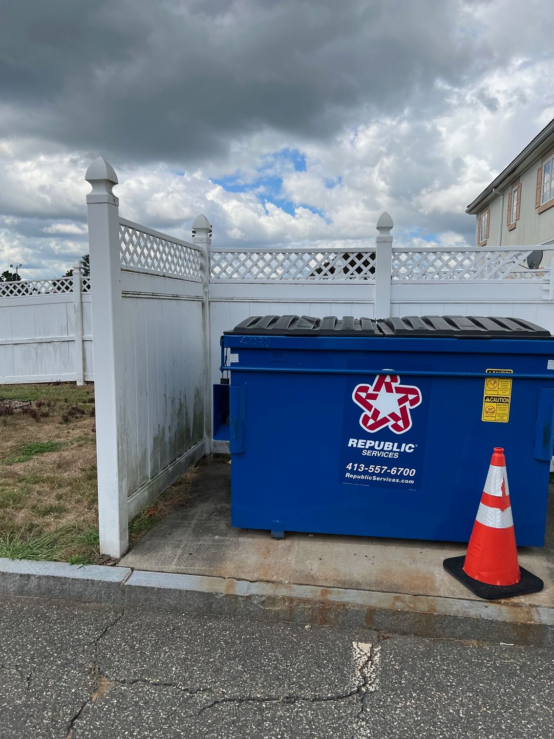A blue dumpster in a fenced enclosure with a traffic cone nearby. The sky is cloudy.