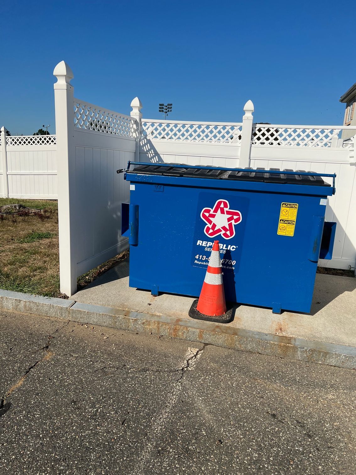 Blue dumpster with a red and white logo, orange cone, white fence, asphalt.
