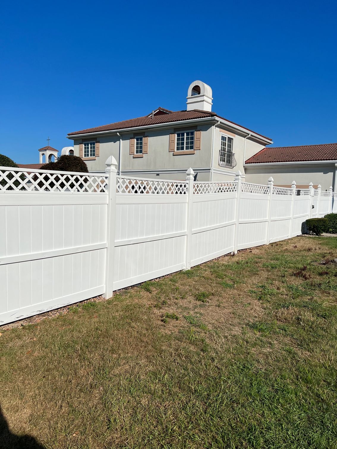White vinyl fence in front of a two-story house with stucco walls, red roof, and chimney under a clear blue sky.