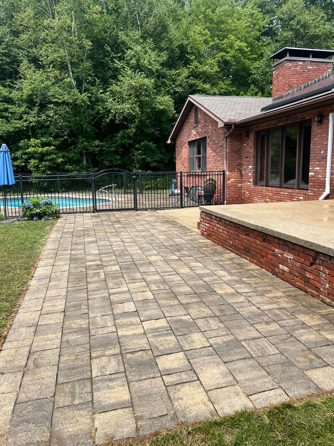 Brick paver patio next to a brick house with a swimming pool in the background and a green yard.