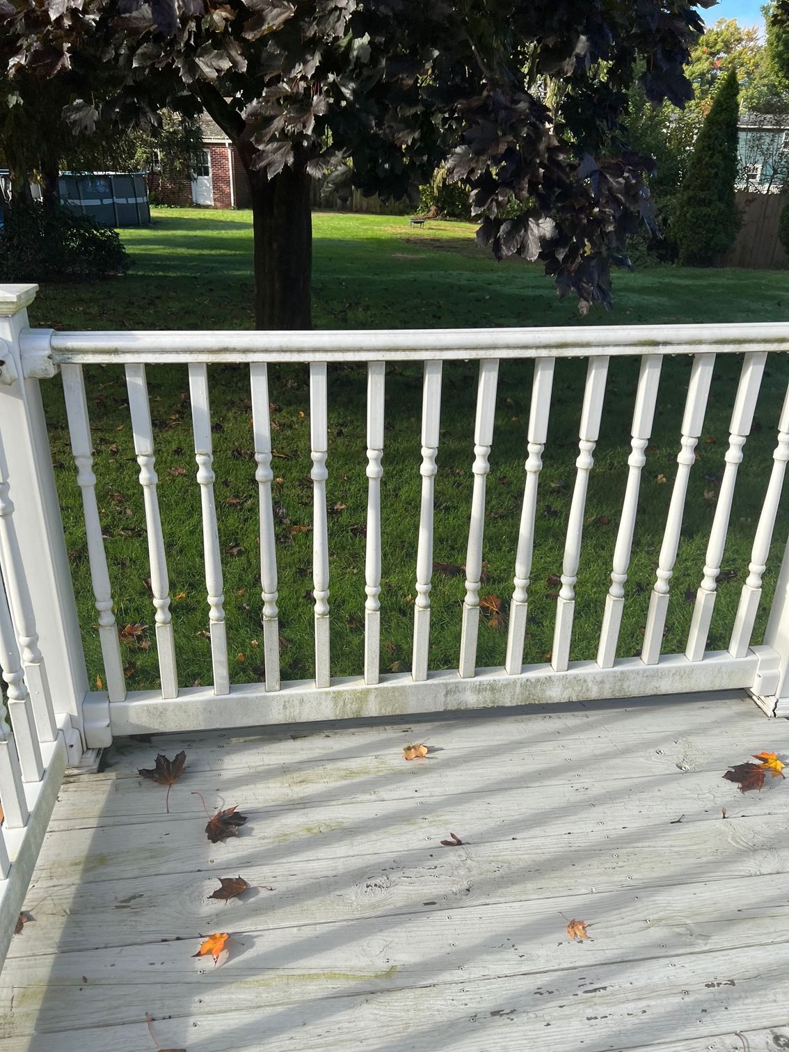 White porch railing with balusters, fallen leaves on the deck, green lawn, tree, and sunny sky.