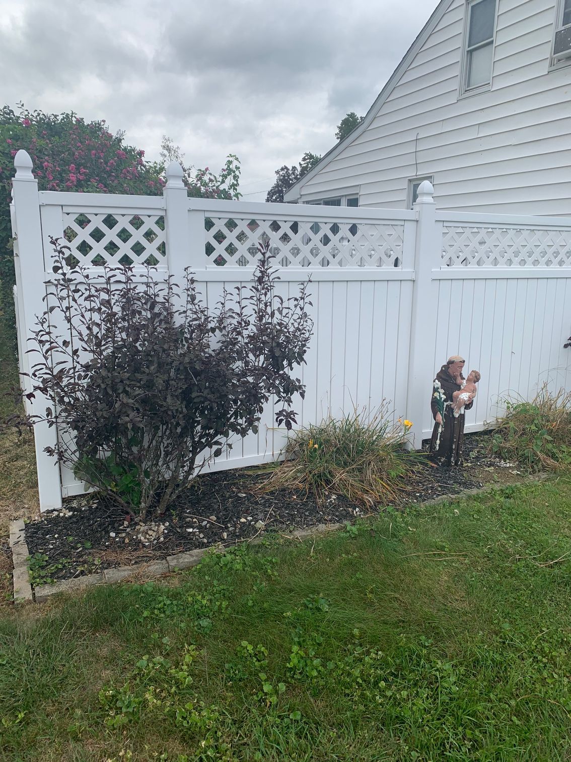 White picket fence with decorative lattice, beside a dark-leaved bush and a garden gnome, on a cloudy day.