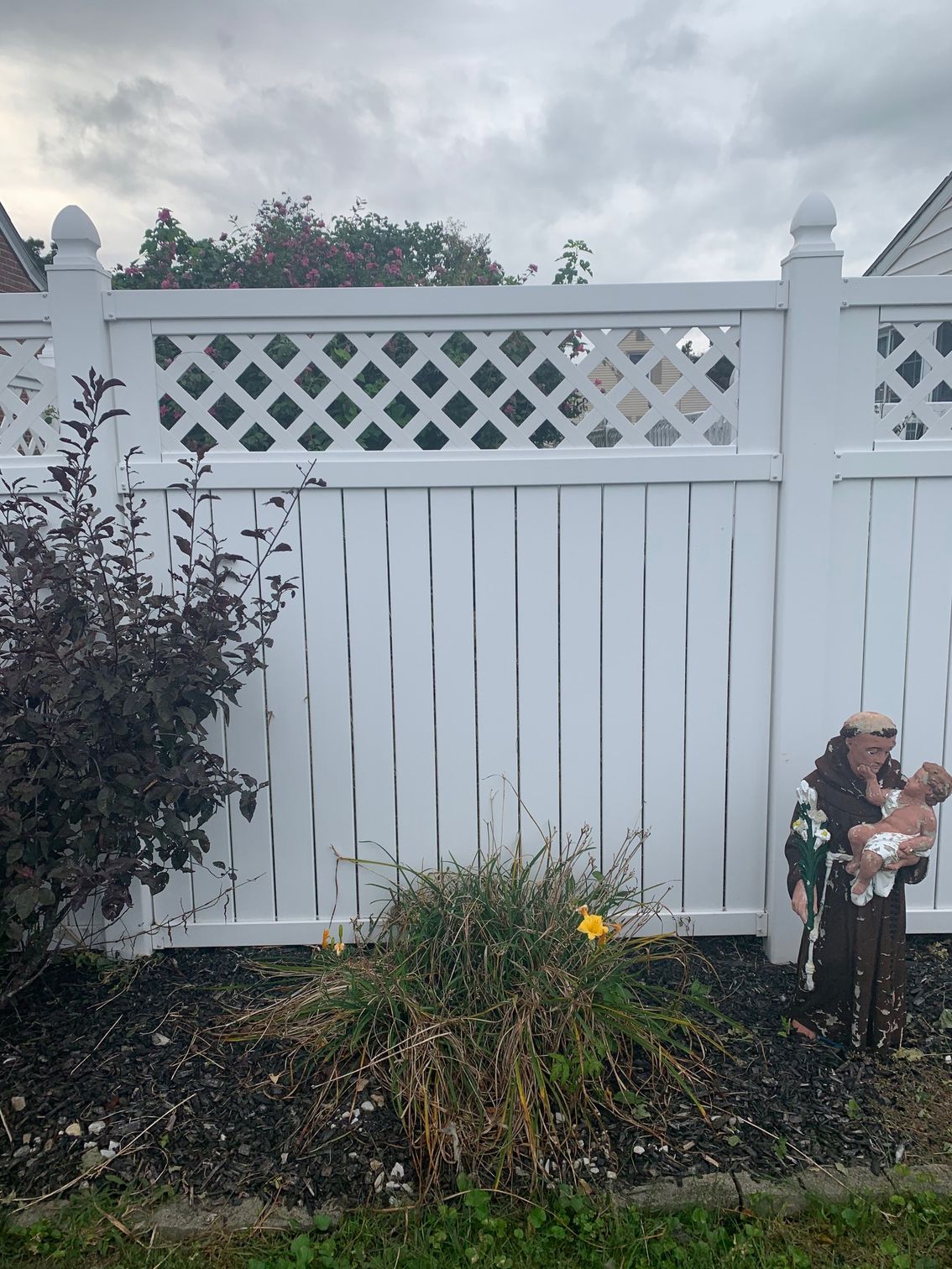 White fence with a lattice top, a statue of an elderly figure holding a baby, and dark mulch with plants in front.