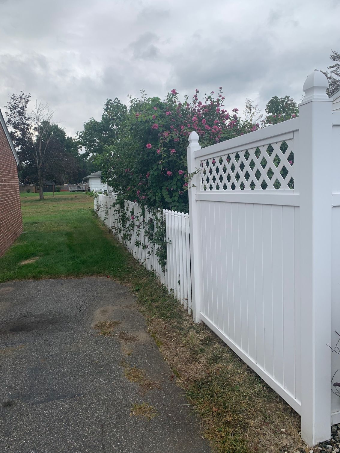 White vinyl fence alongside a driveway and a grassy area. In the background are trees and a cloudy sky.