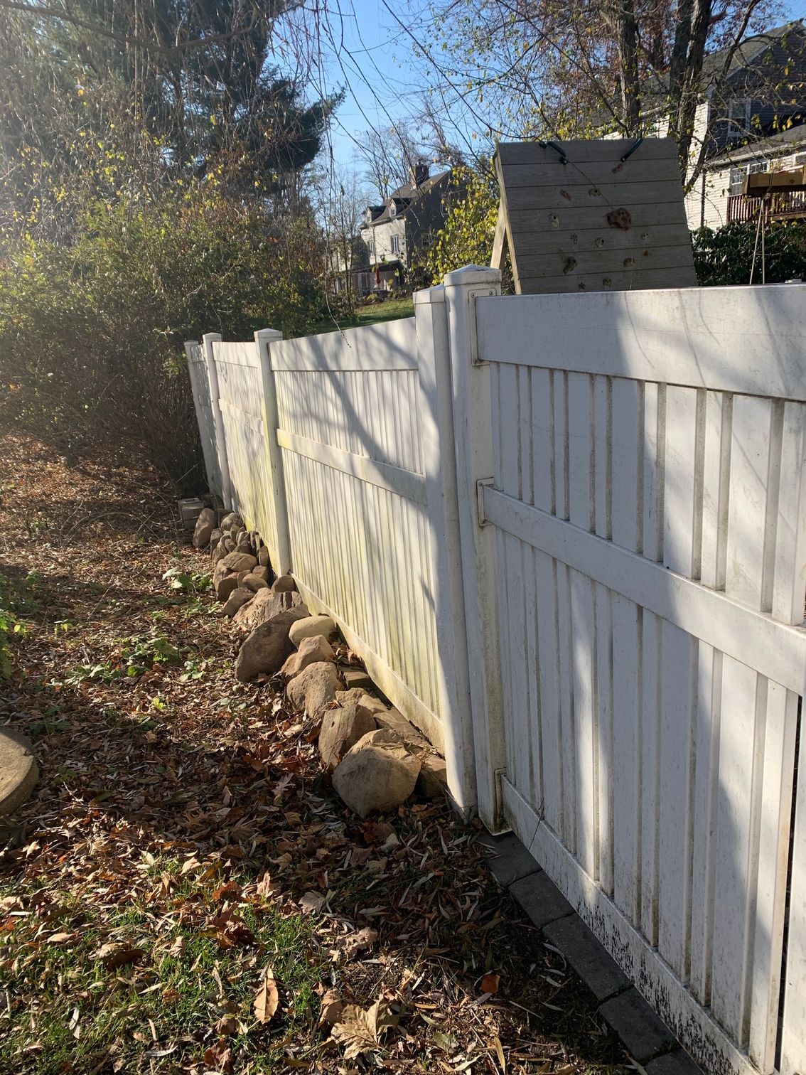 White fence with several large rocks along its base, in a yard with fallen leaves. Sunlight casts shadows.