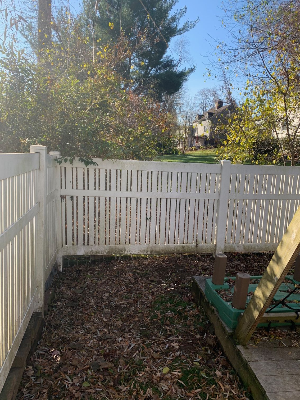 White picket fence surrounds a yard with leaves and a green play set, trees and houses in the background.